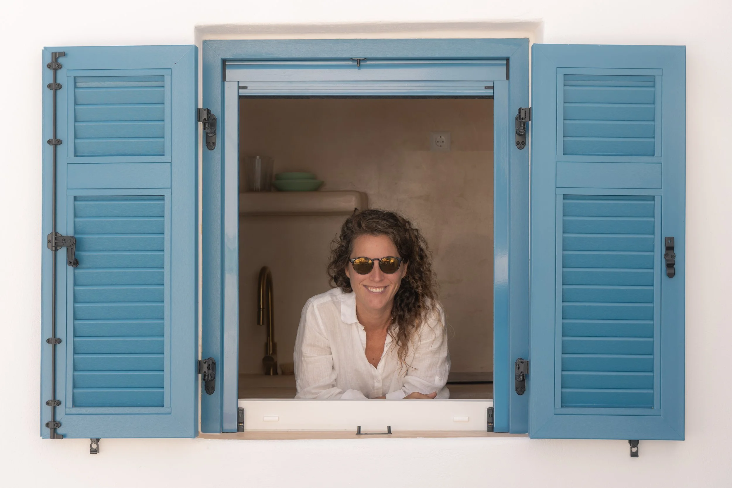 Smiling woman with curly hair and sunglasses leaning out of blue window with shutters.