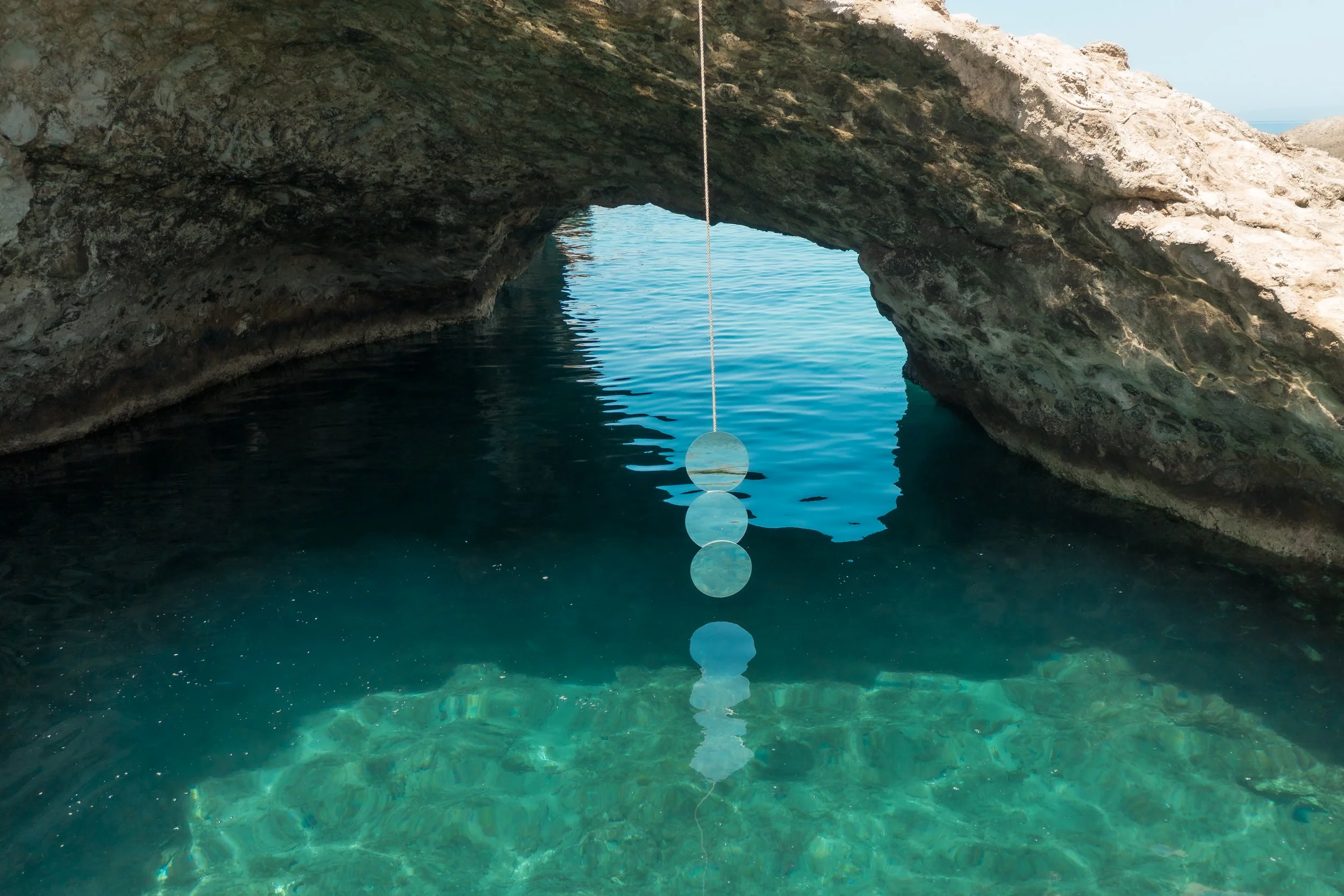 Natural rock formation with an arched opening over a body of clear, turquoise water, reflecting the sky and rocks.