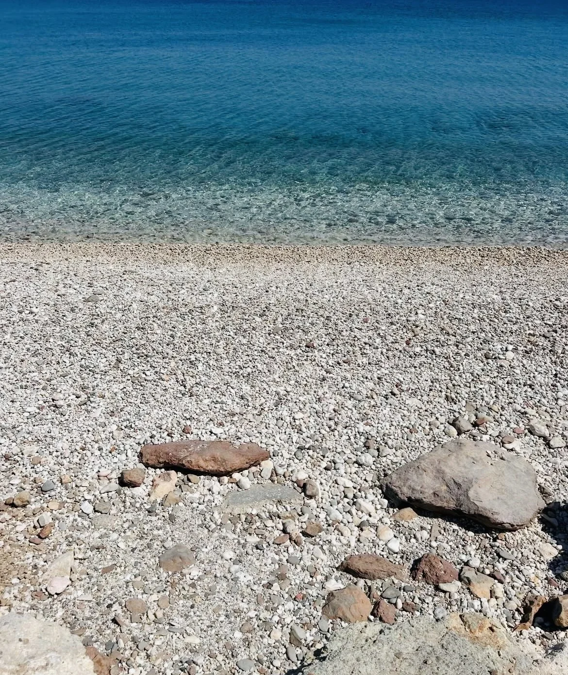 Pebble beach with large rocks near the shoreline, clear blue water, and gentle waves in the background.