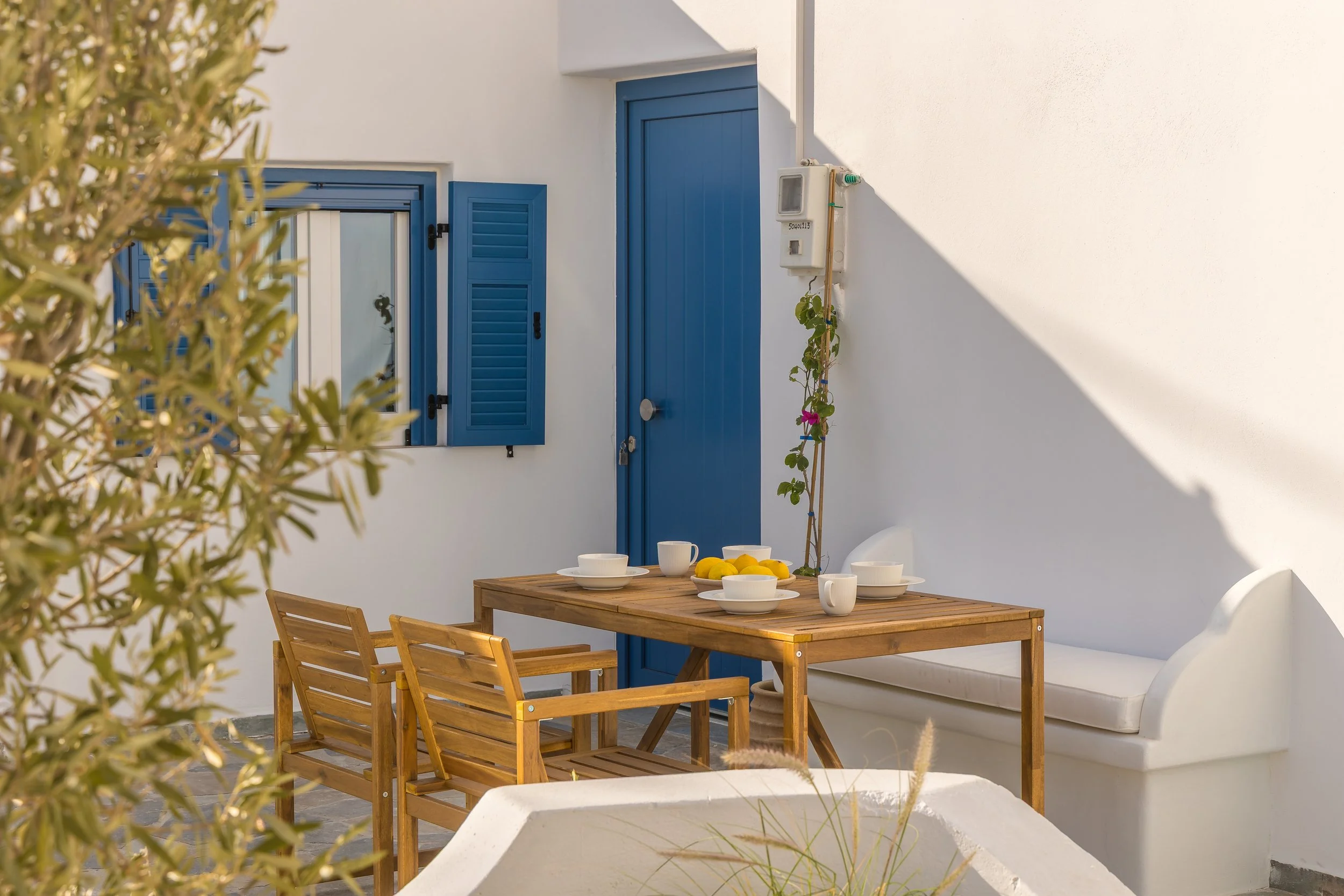 Outdoor dining area with a wooden table set with white bowls, cups, and lemons, surrounded by wooden chairs, against a white wall with blue doors and shutters, and greenery in the foreground.
