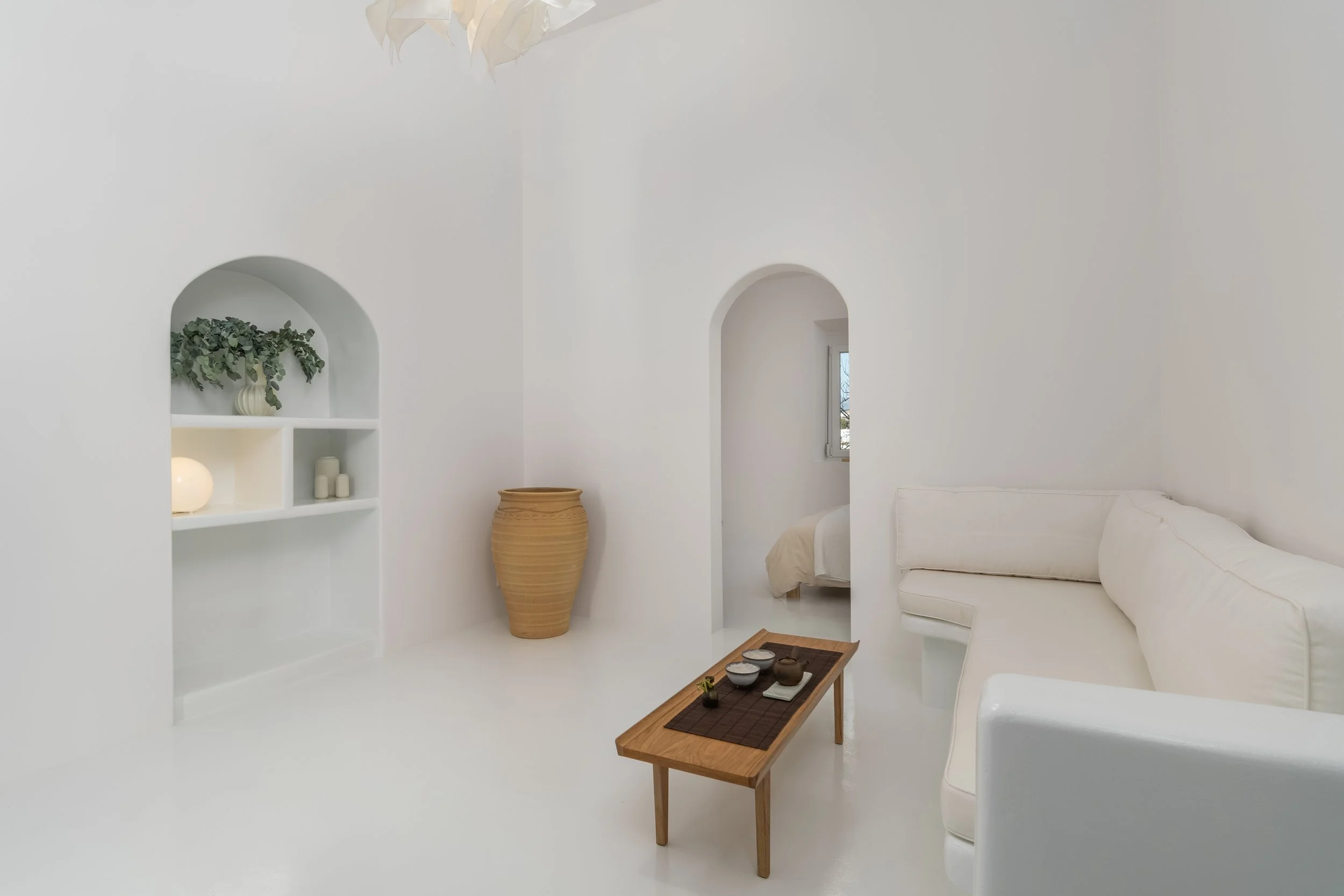 Minimalist white living room with a built-in arched shelf, potted green plant, large beige vase, white sofa, small wooden coffee table with candles, small window, and modern white decor.