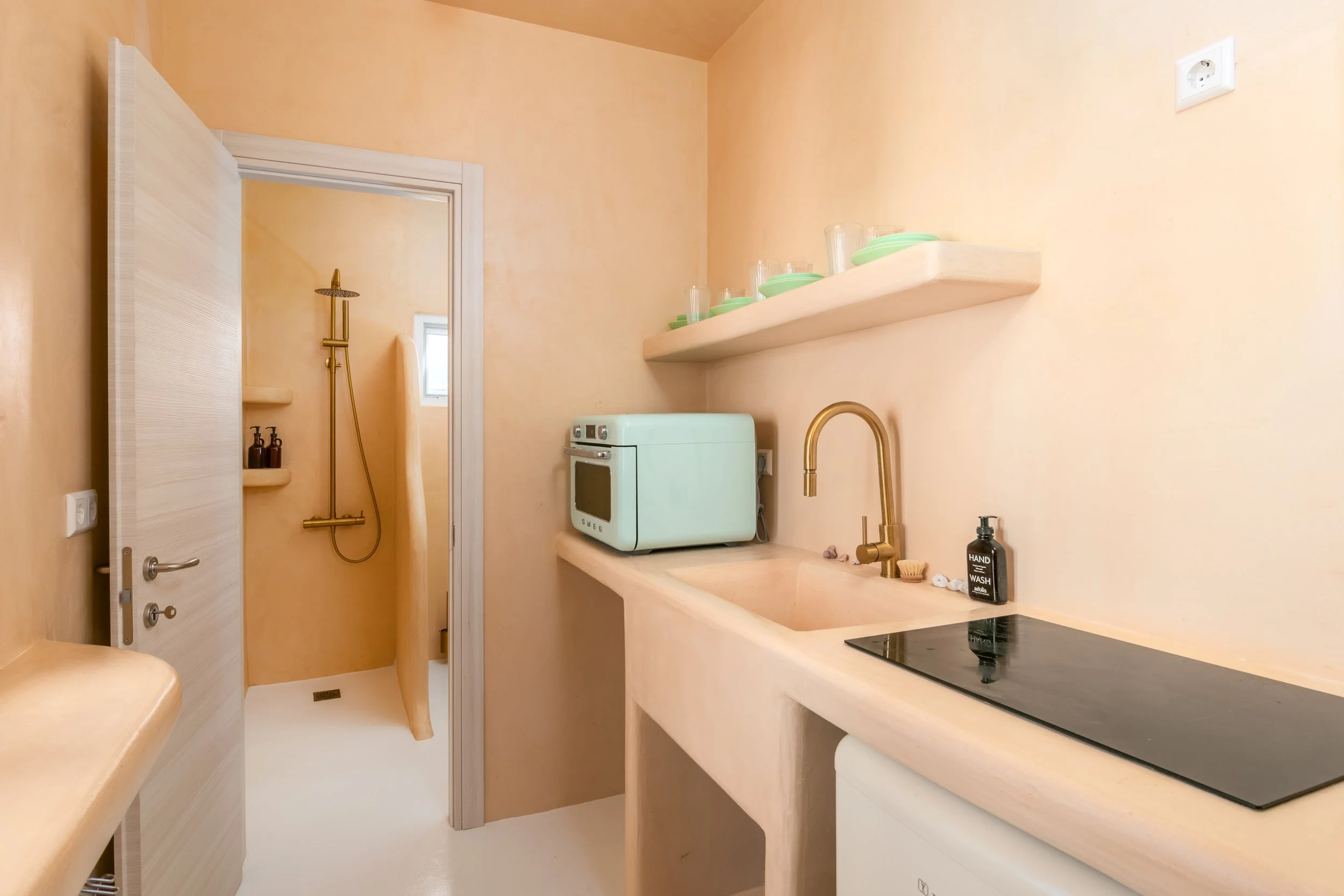 A small, minimalist kitchen with peach-colored walls and a beige countertop. There is a small stovetop, a black soap or lotion dispenser, a gold faucet, a white countertop oven, green dishes, and glasses on a shelf. A window is visible in the shower area, which also has a gold showerhead and handheld shower. The area features a small open space with a door leading to the shower.