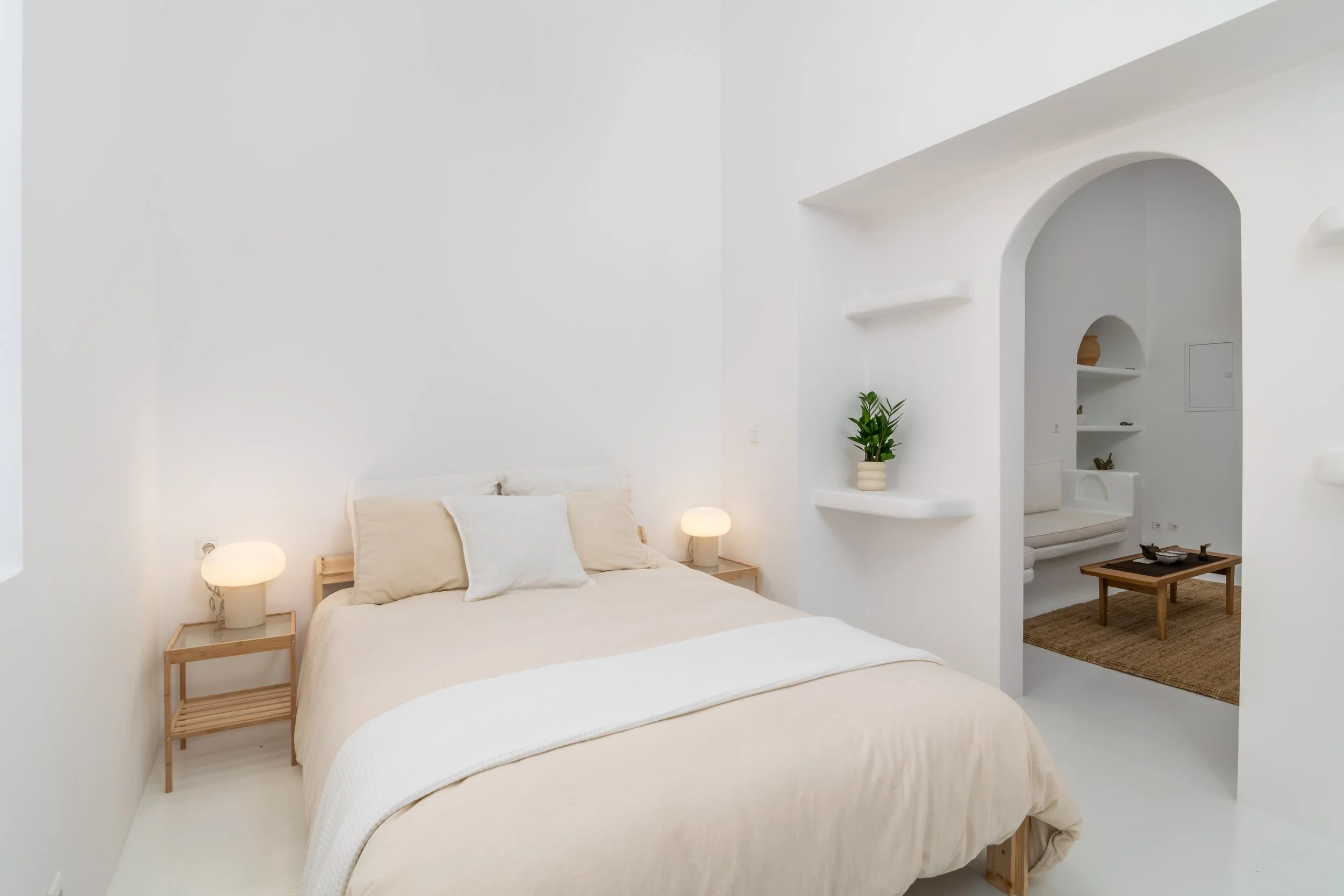 A minimalist bedroom with white walls and bedding, a wooden nightstand with a lamp, and built-in shelves with a plant and decorative items. An arch leads into another room with a small table and seating area.