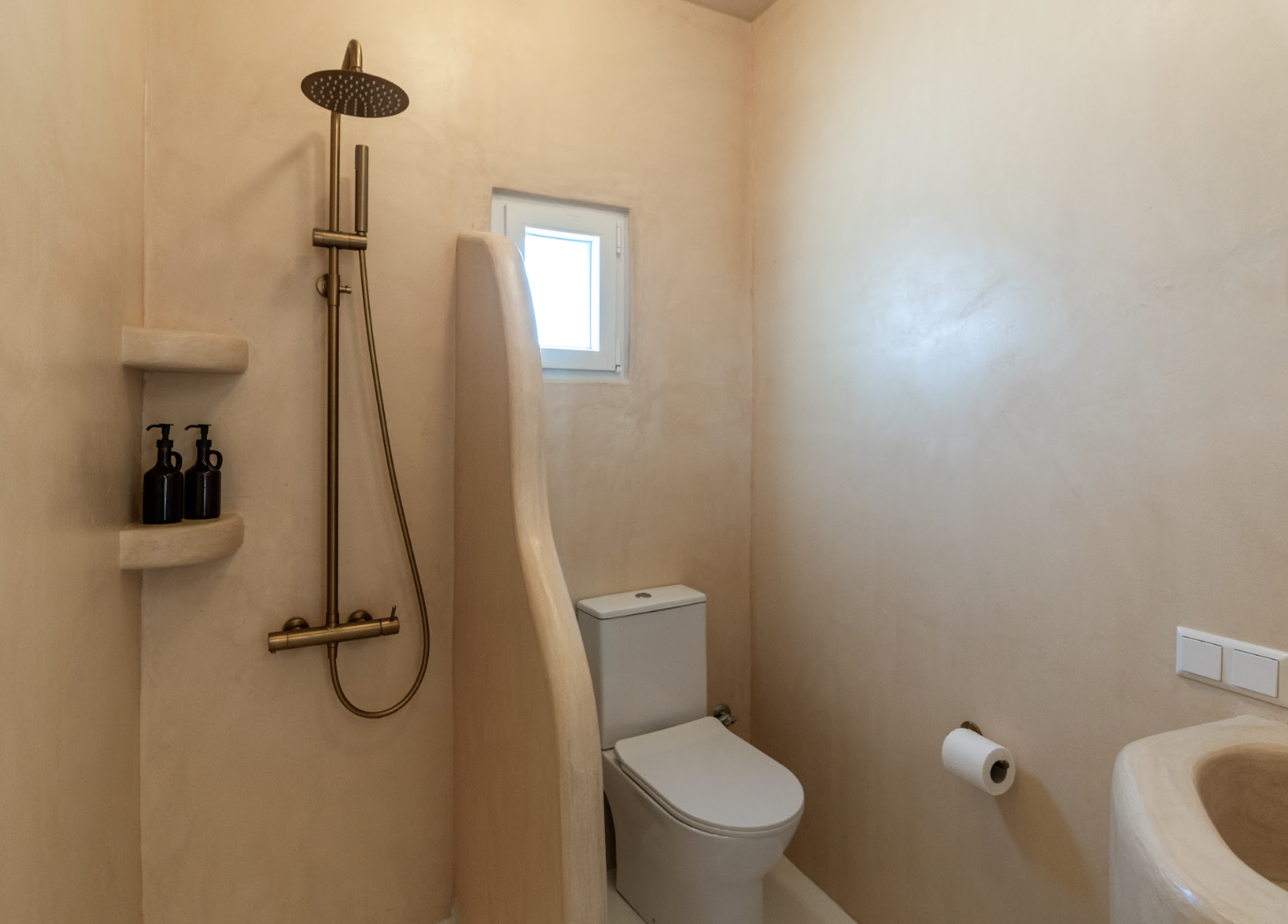 Small bathroom with beige walls, a window, a toilet, a shower with a gold shower head, a built-in shelf with two black pump bottles, and a wall-mounted toilet paper holder.