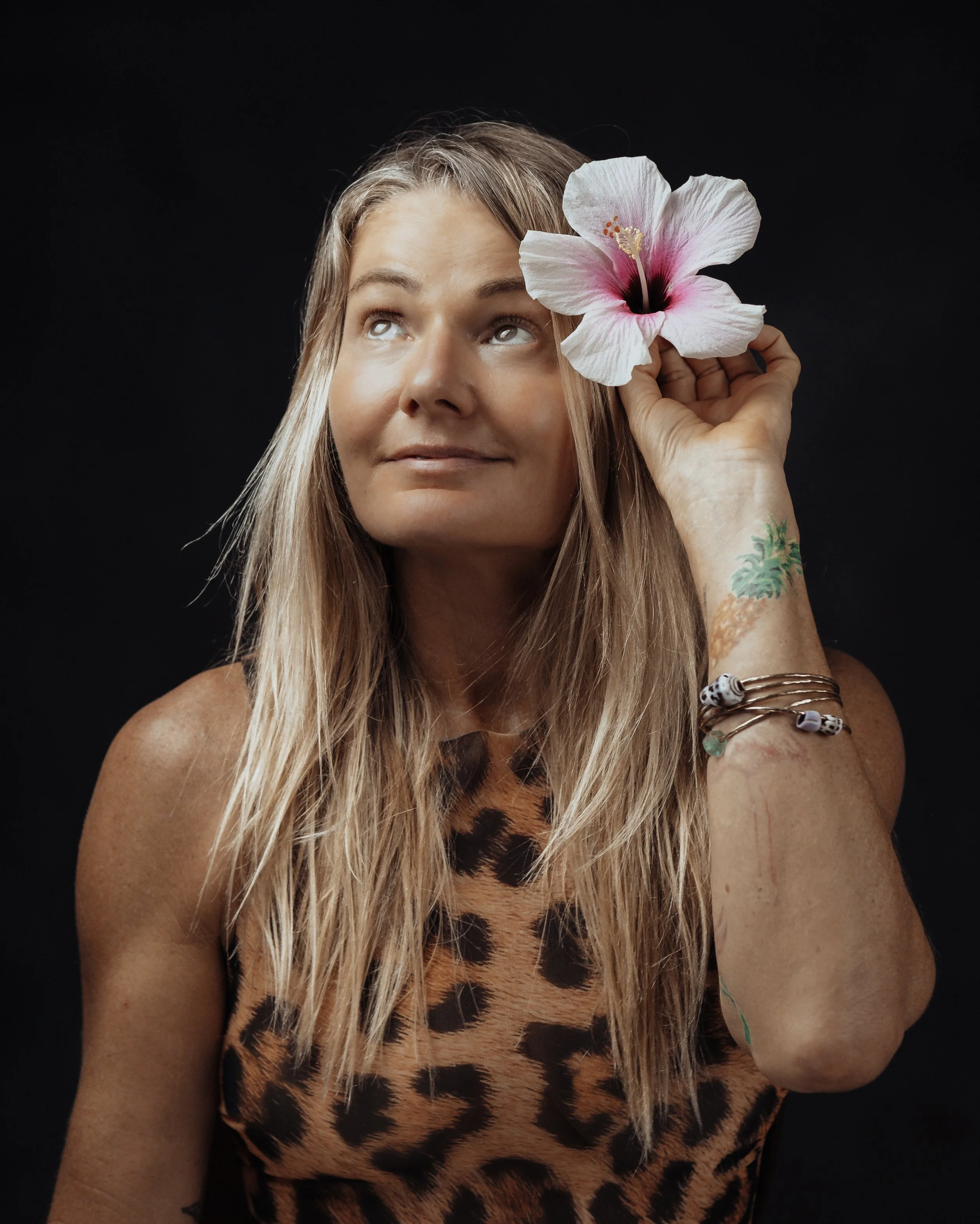 A woman with long blonde hair holding a large white and pink hibiscus flower near her temple, wearing a leopard print sleeveless top, with tattoos and bracelets on her left arm, against a black background.