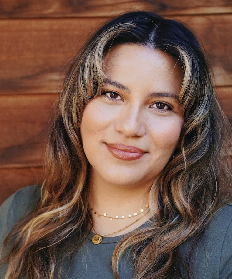 Close-up portrait of a woman with wavy brown hair and light highlights, smiling softly in front of a wooden background.