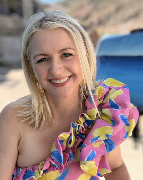 A woman with blonde hair smiling at the camera, wearing a colorful floral dress with ruffles on one shoulder, outdoors near a beach.