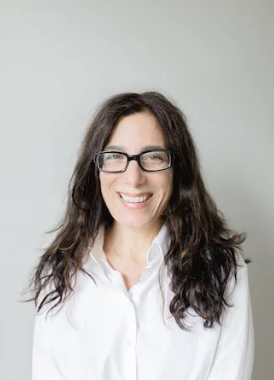 A woman with dark brown hair and glasses smiling, wearing a white blouse, positioned against a plain light gray background.
