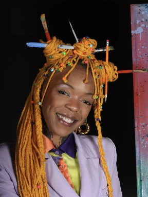 A woman with yellow dreadlocks decorated with colorful beads and pencils, smiling in front of a dark background.