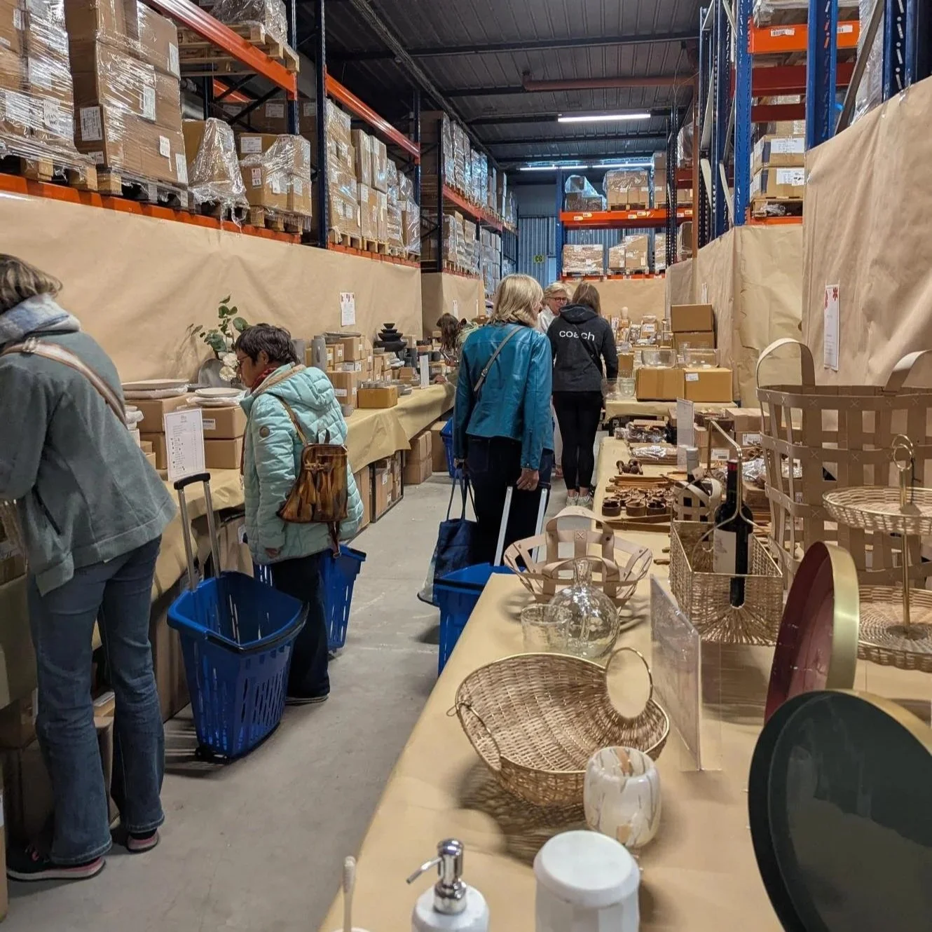 People shopping at a warehouse-style store with shelving filled with boxed items, tables displaying various home decor and cookware, and shopping carts lined up along the aisle.