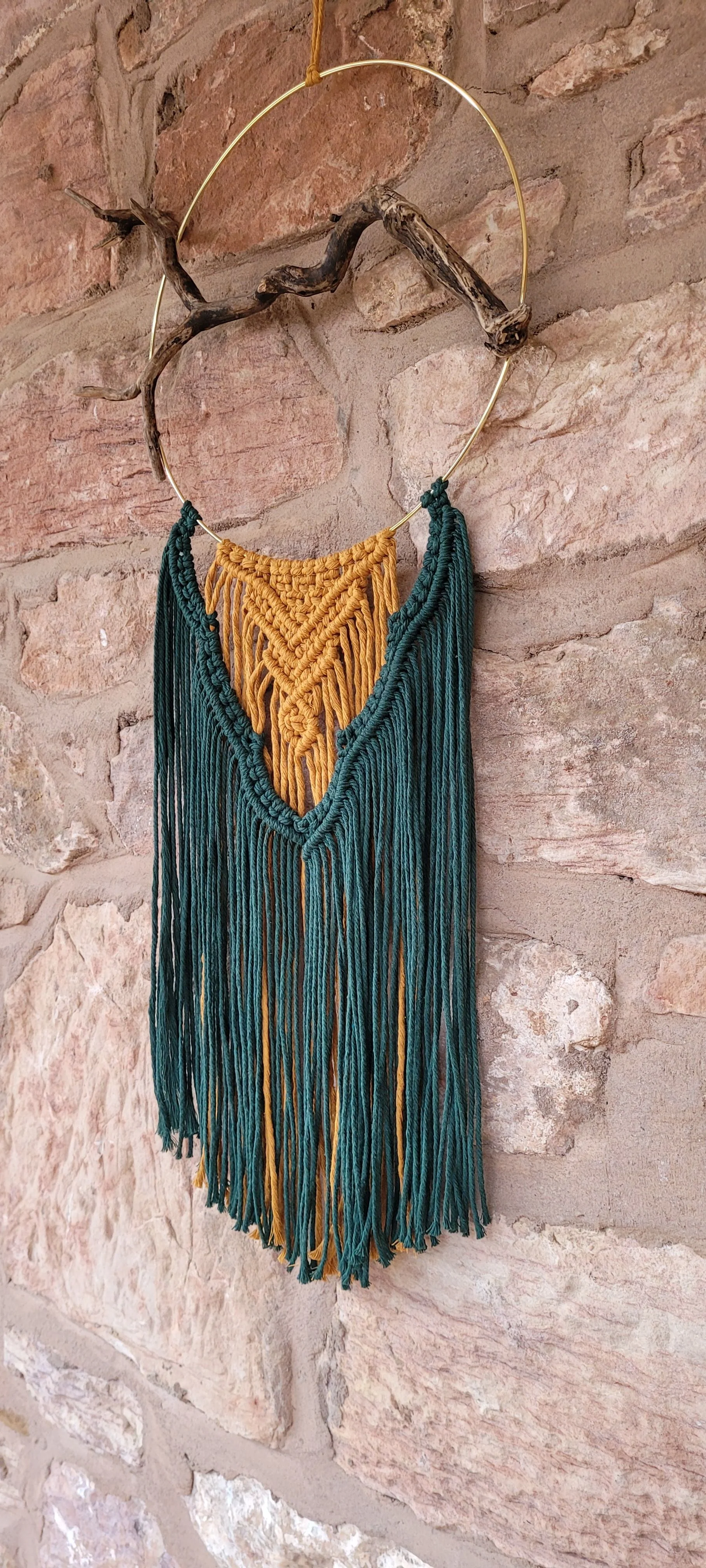 A decorative wall hanging crafted with green and yellow yarn, featuring a macrame design, hangs from a golden circular frame attached to a brick wall.