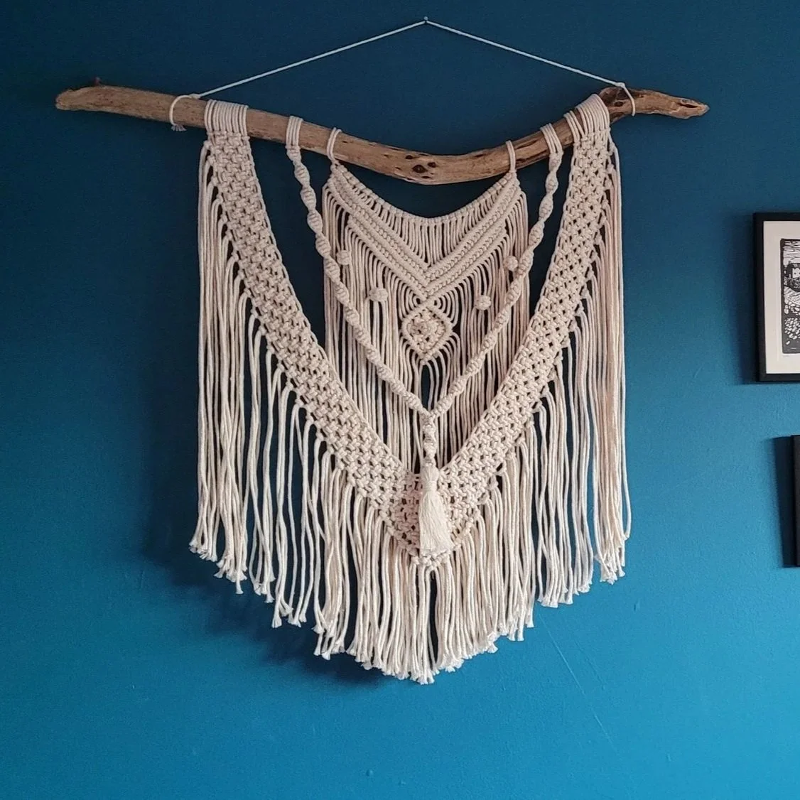 Cream-colored macramé wall hanging with intricate patterns, hanging from a driftwood rod on a teal wall.