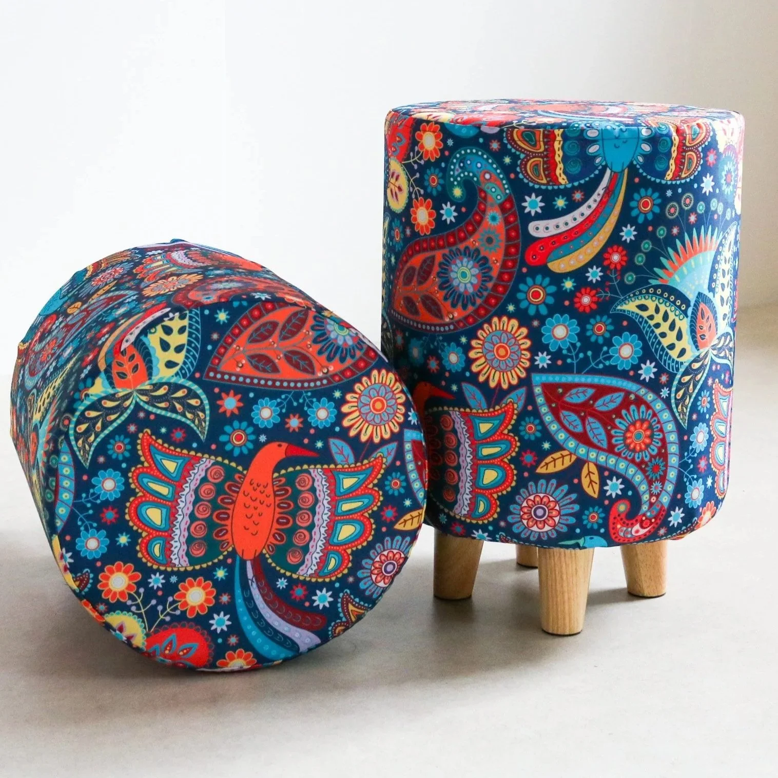 Two pieces of furniture with bright, colorful abstract floral and paisley patterns on a white background, one is a rounded ottoman and the other is a cubic stool with wooden legs.