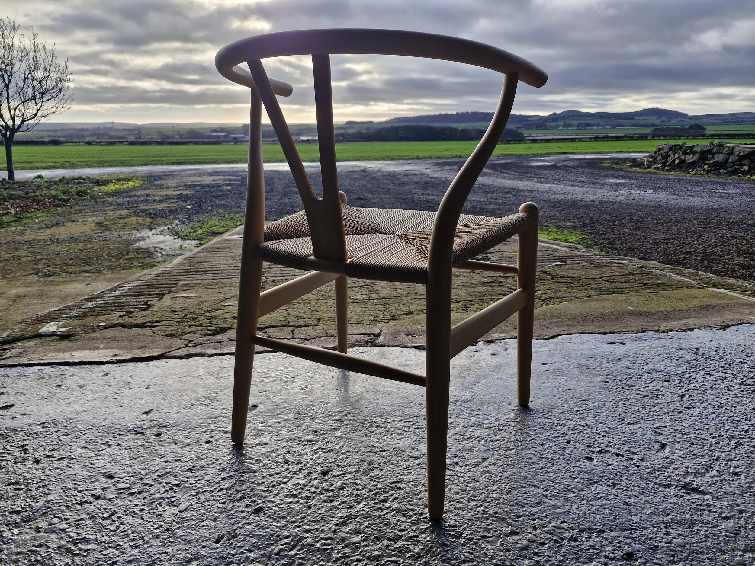 A wooden chair on a weathered stone porch overlooking a rural landscape with green fields, a winding road, and rolling hills under cloudy skies.