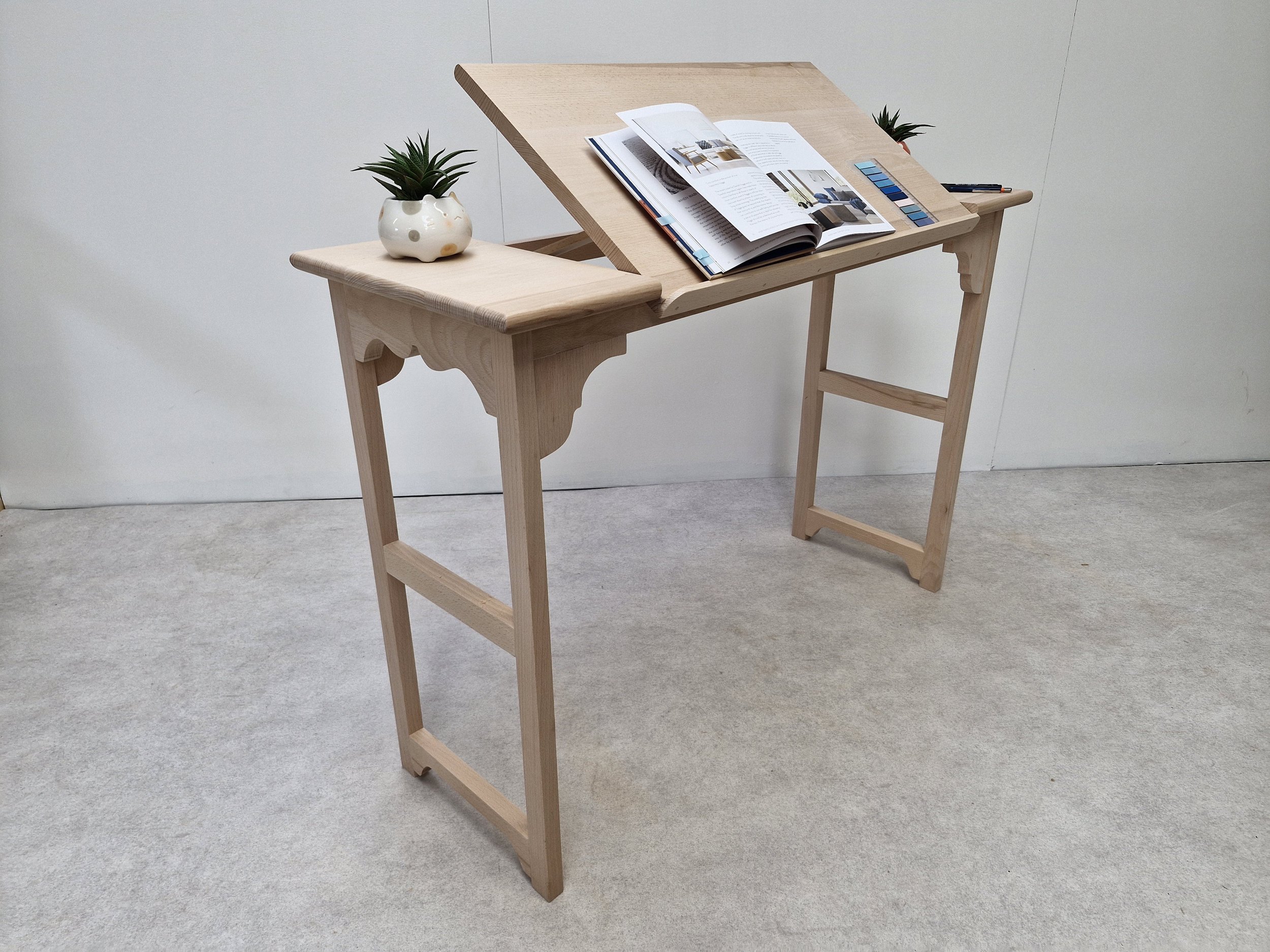 Wooden drafting table with an open magazine, color swatches, and pens on a blank wall background.