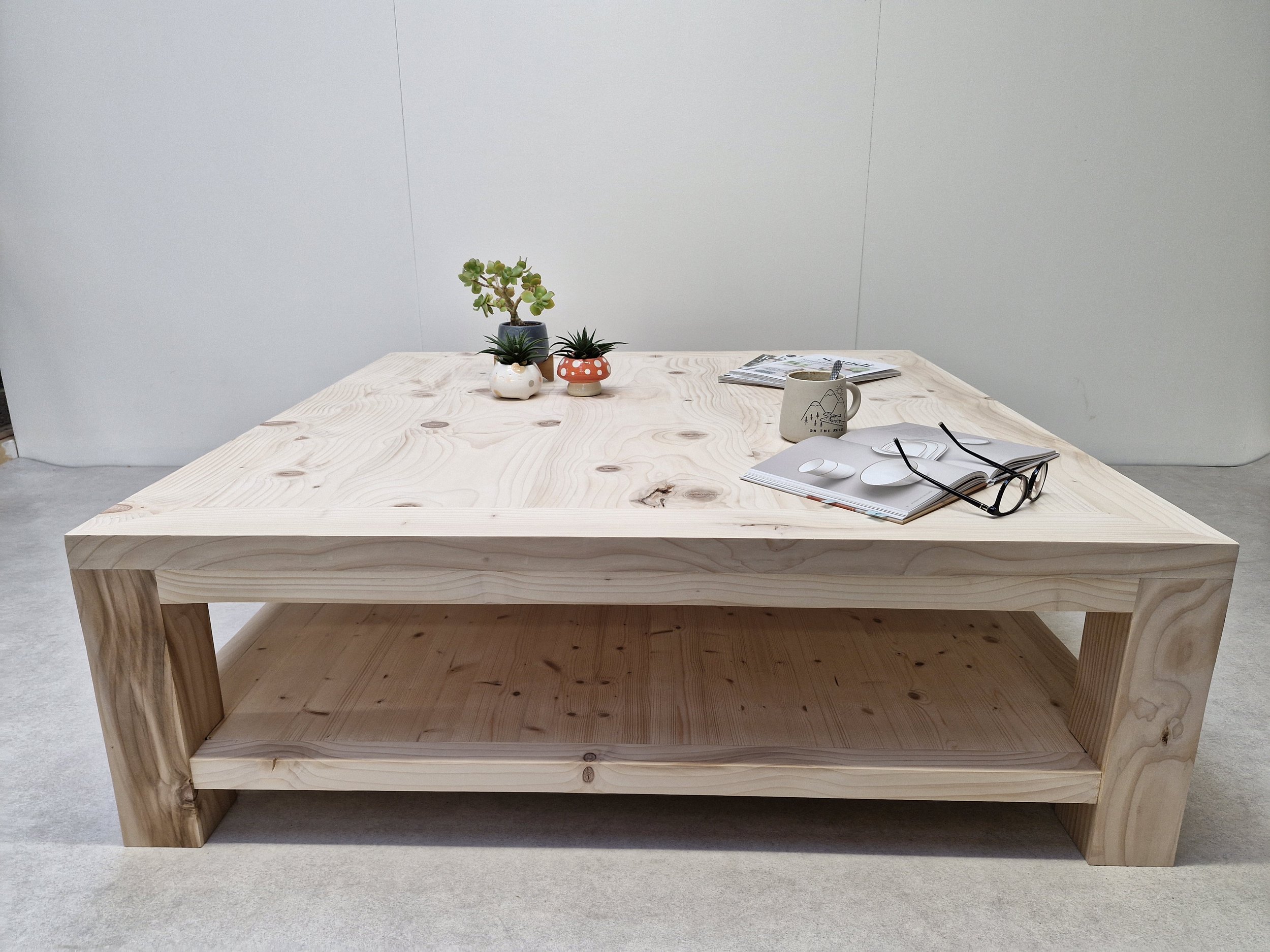 A light-colored wooden table with potted plants, a book, glasses, a coffee mug, and a pair of earbuds is shown against a plain, light-colored wall.