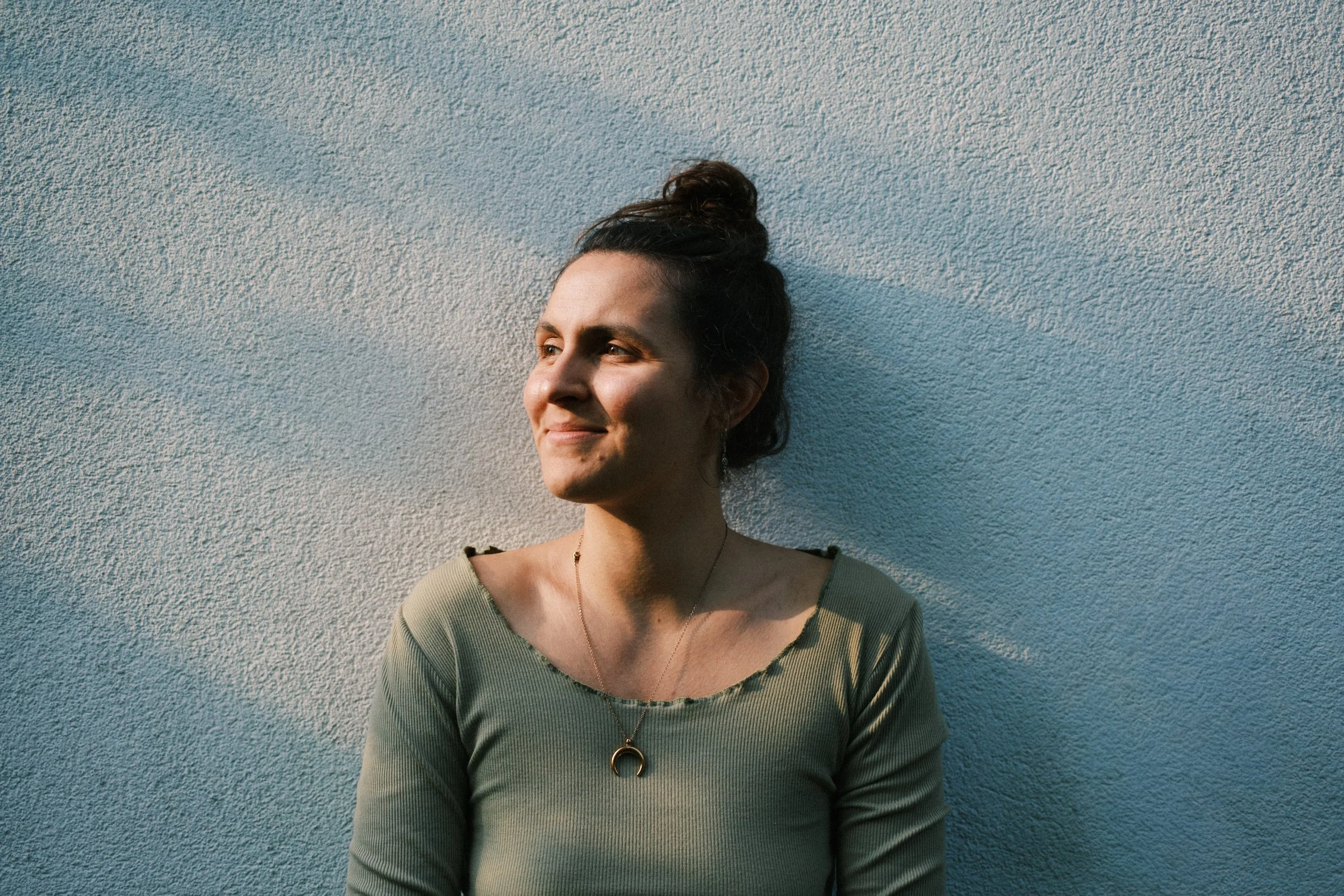 Junge Frau mit dunklem Haar in einem hohen Dutt, lächelnd, steht vor einer hellblauen Wand, trägt ein grünes Oberteil und eine Halskette mit einem Halbmonden-Anhänger.