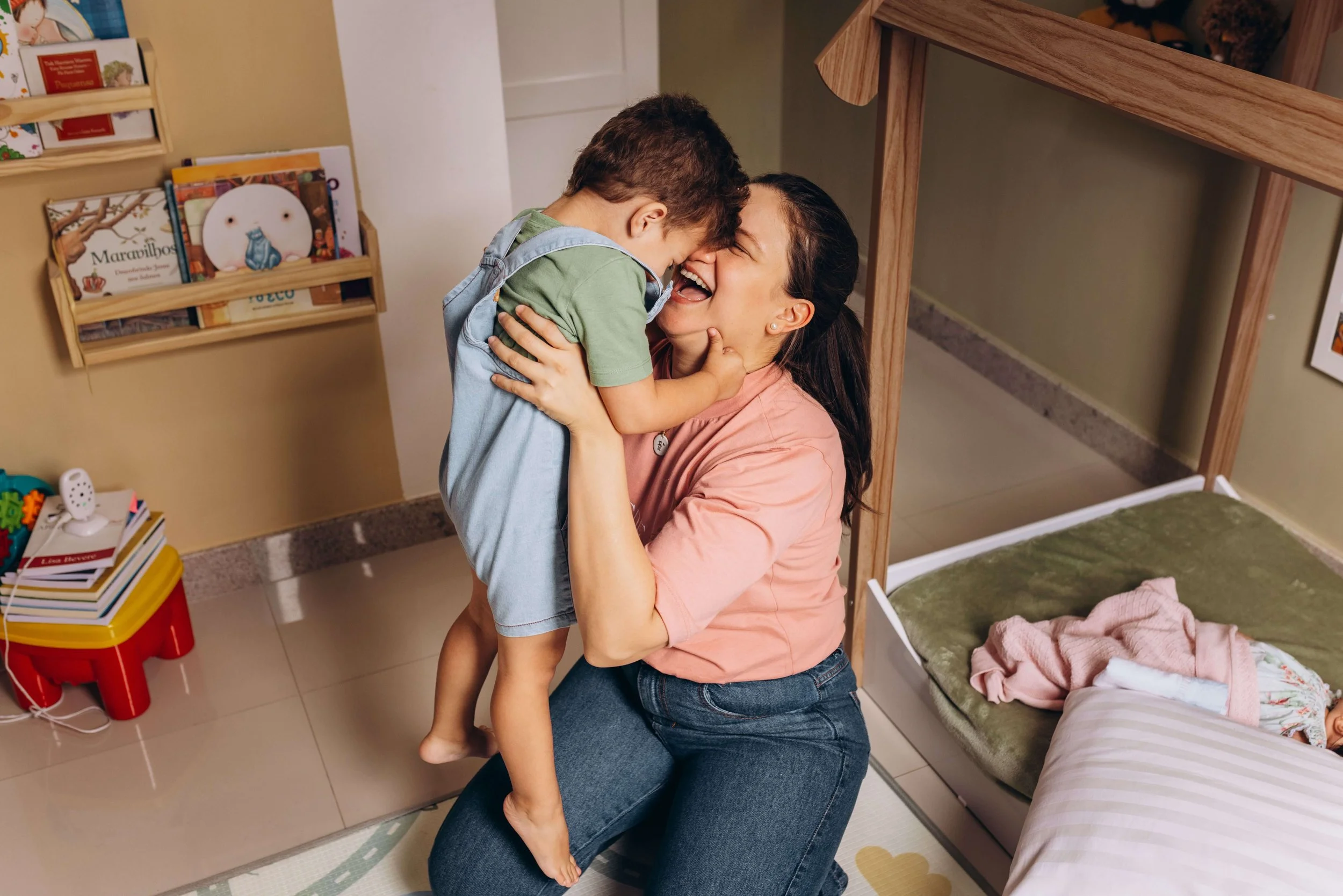 A parent and young child in Tumwater, WA, connecting and laughing on the floor during in-home speech therapy. Mom is sitting on the floor holding the boy up in the air. They are laughing and touching foreheads. The room has various toys and books.