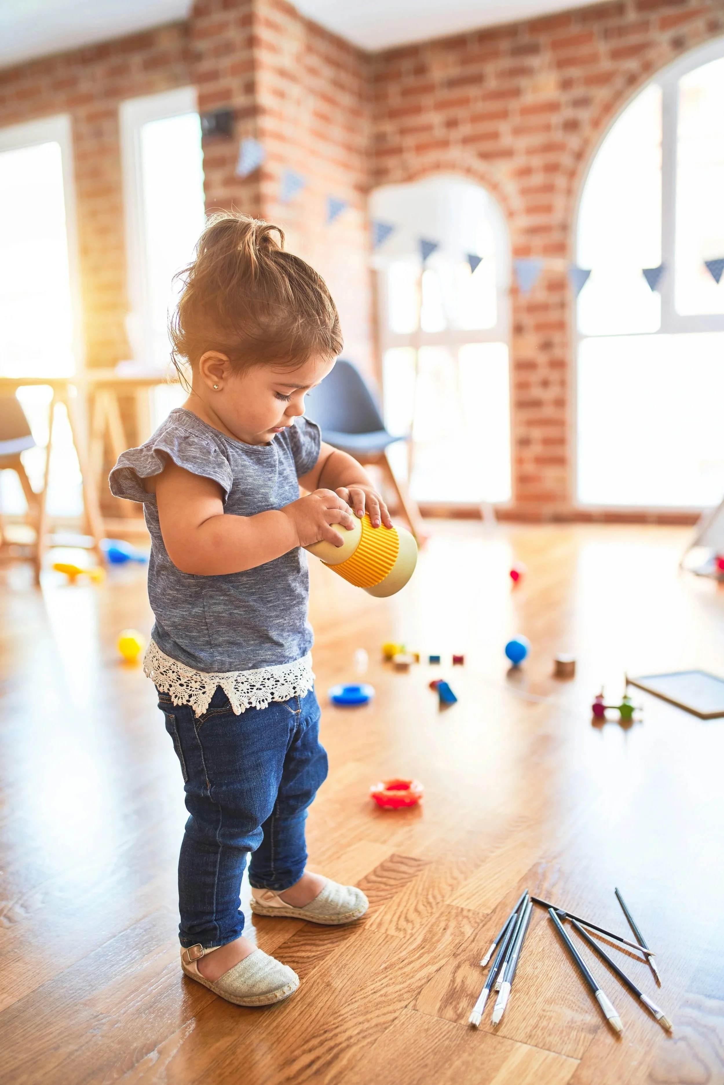 Young girl playing with building sticks on wooden floor in a bright room with brick walls and large windows.