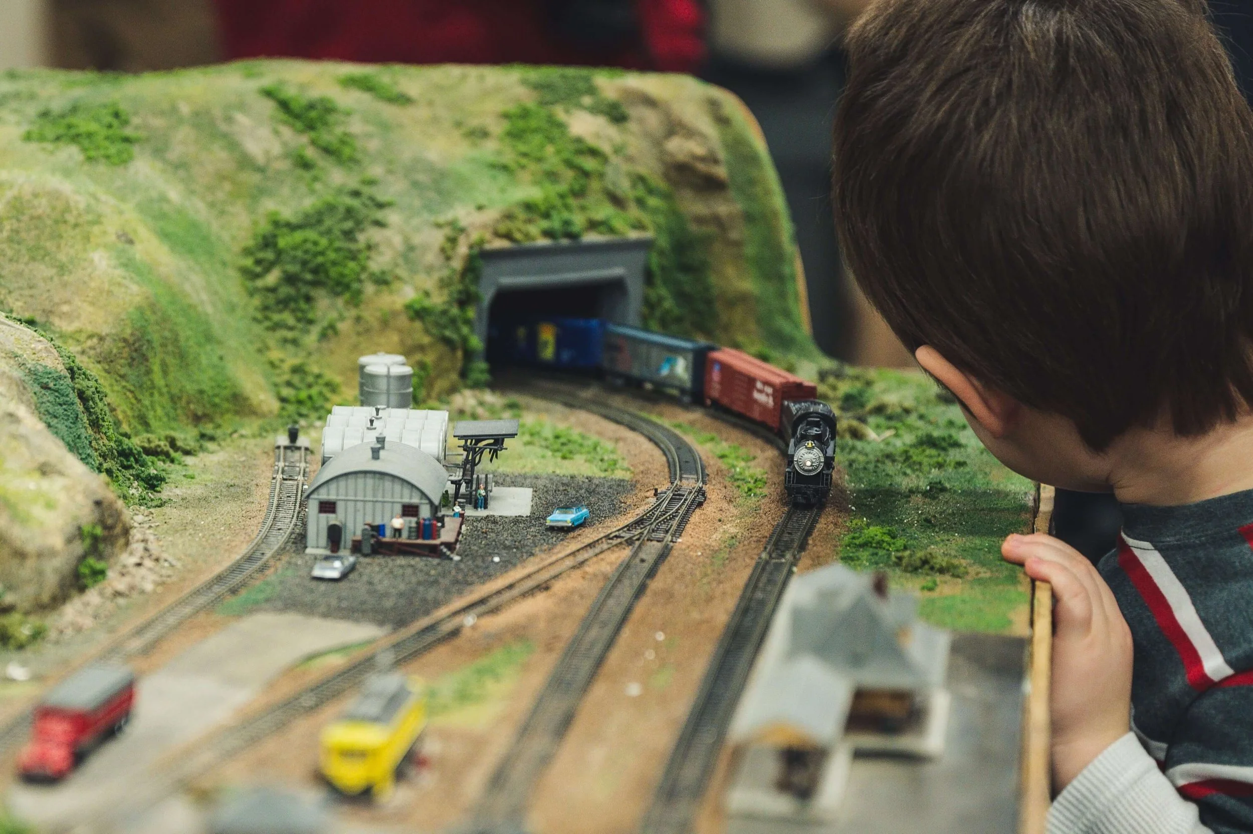 The back of a child's head as he plays by observing and studying toys closely. Here he looks at a toy train as it goes past miniature buildings and along tracks into a tunnel that goes through a miniature grassy hill.