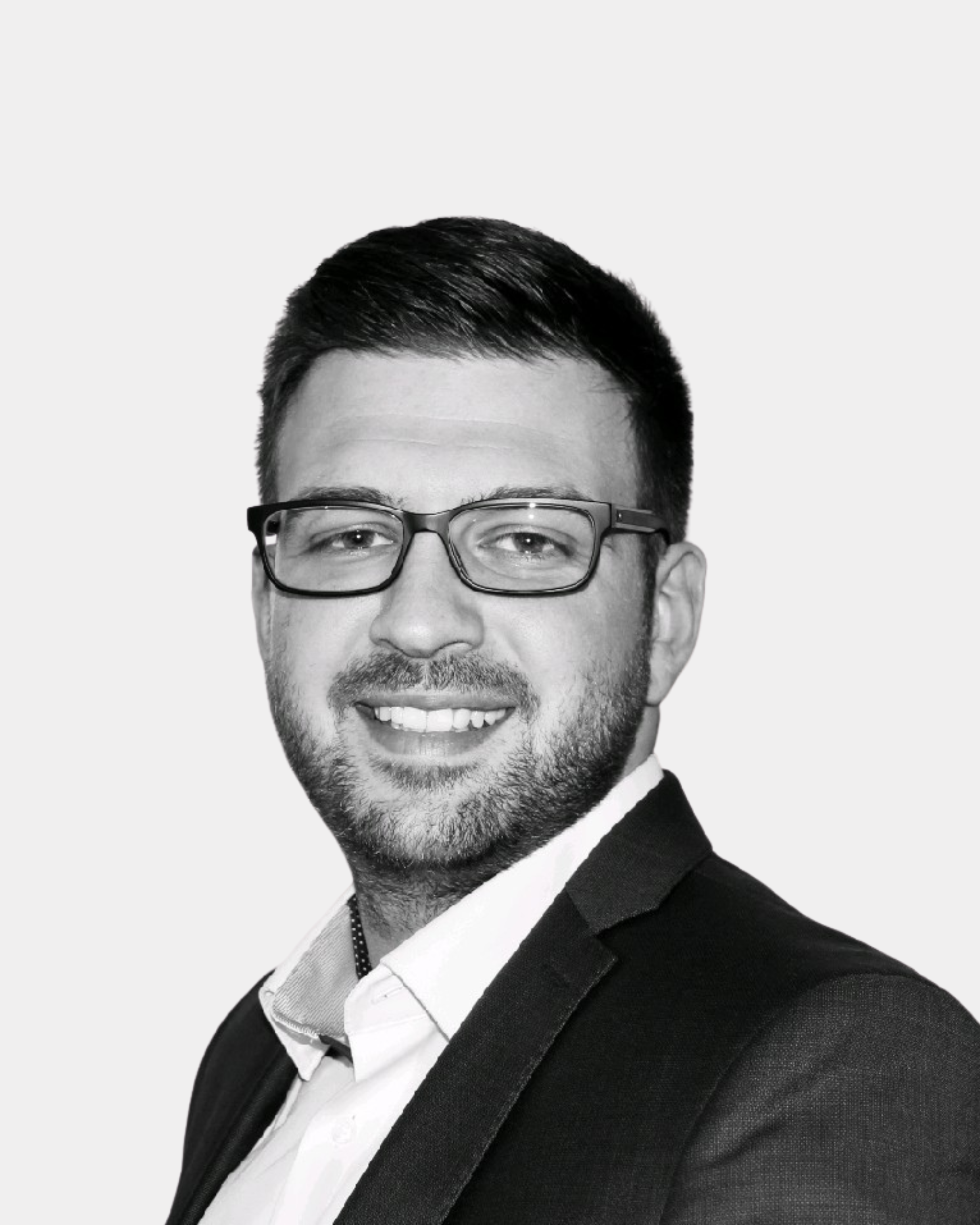 Schwarz-weiß-Bild eines lächelnden Mannes mit Brille, kurzer dunkler Haarfrisur, Anzug, weißem Hemd und Krawatte, gegen weißen Hintergrund.