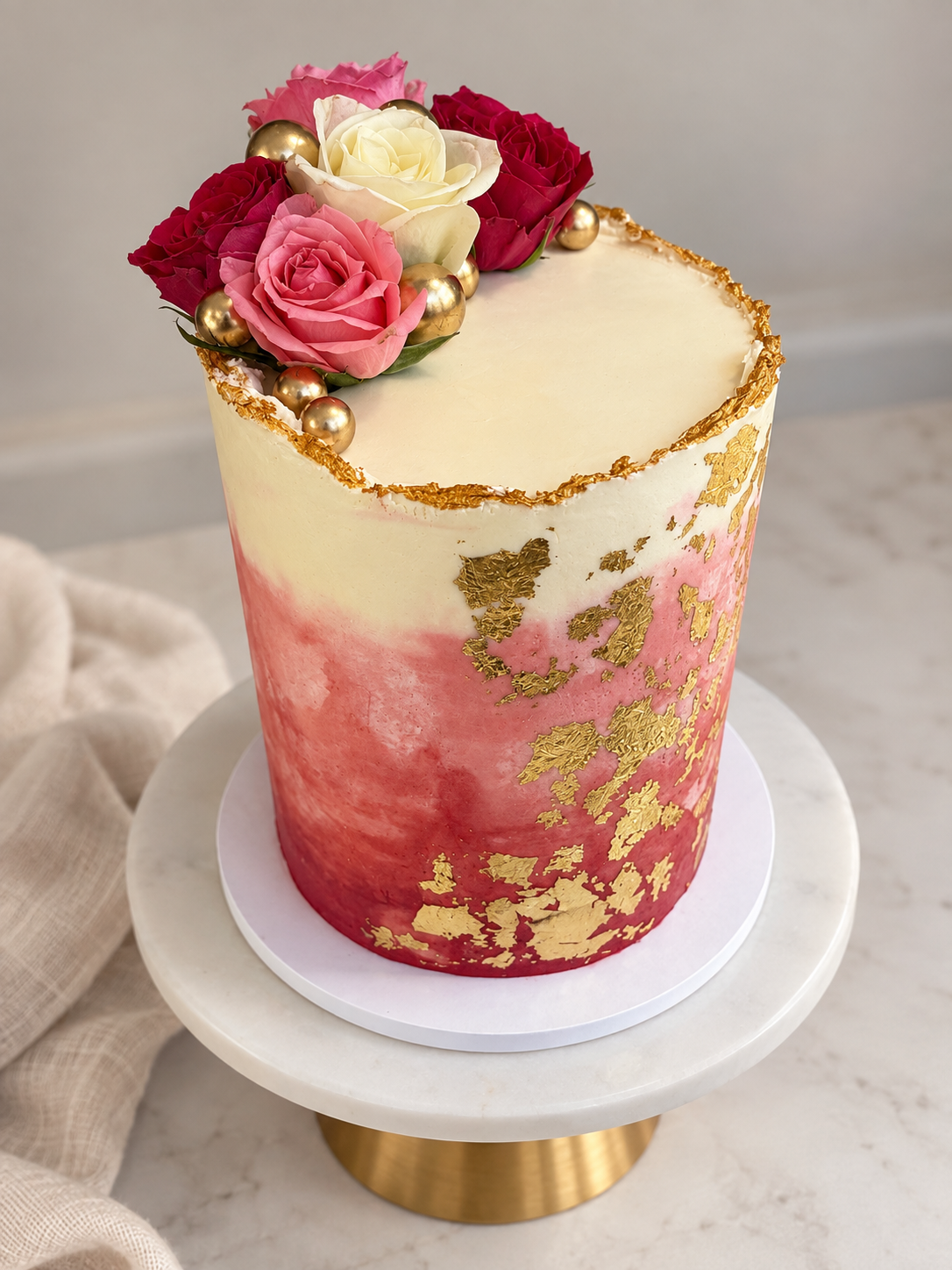 Tall, cylindrical cake with white and pink marbled icing, decorated with gold leaf accents, pink and white roses, and gold balls on top, displayed on a white marble cake stand with a gold base.