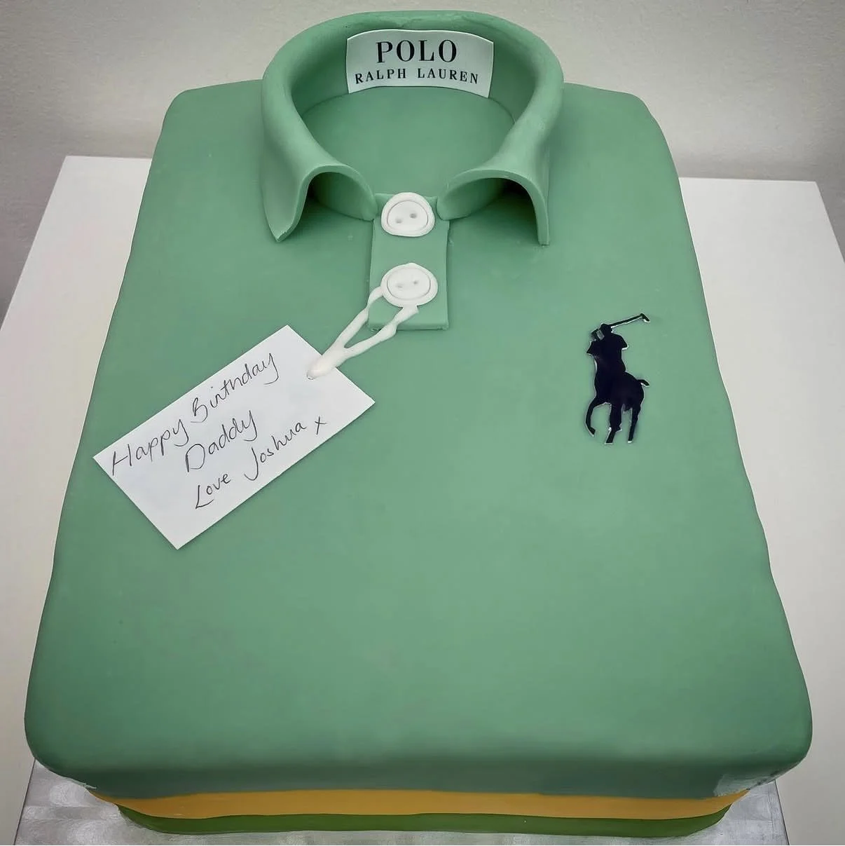 Cake designed to look like a green polo shirt with a collar, buttons, and a label that says "POLO RALPH LAUREN." There is a small black polo player figurine on the side and a birthday note that reads, "Happy Birthday Daddy Love Joshua x."