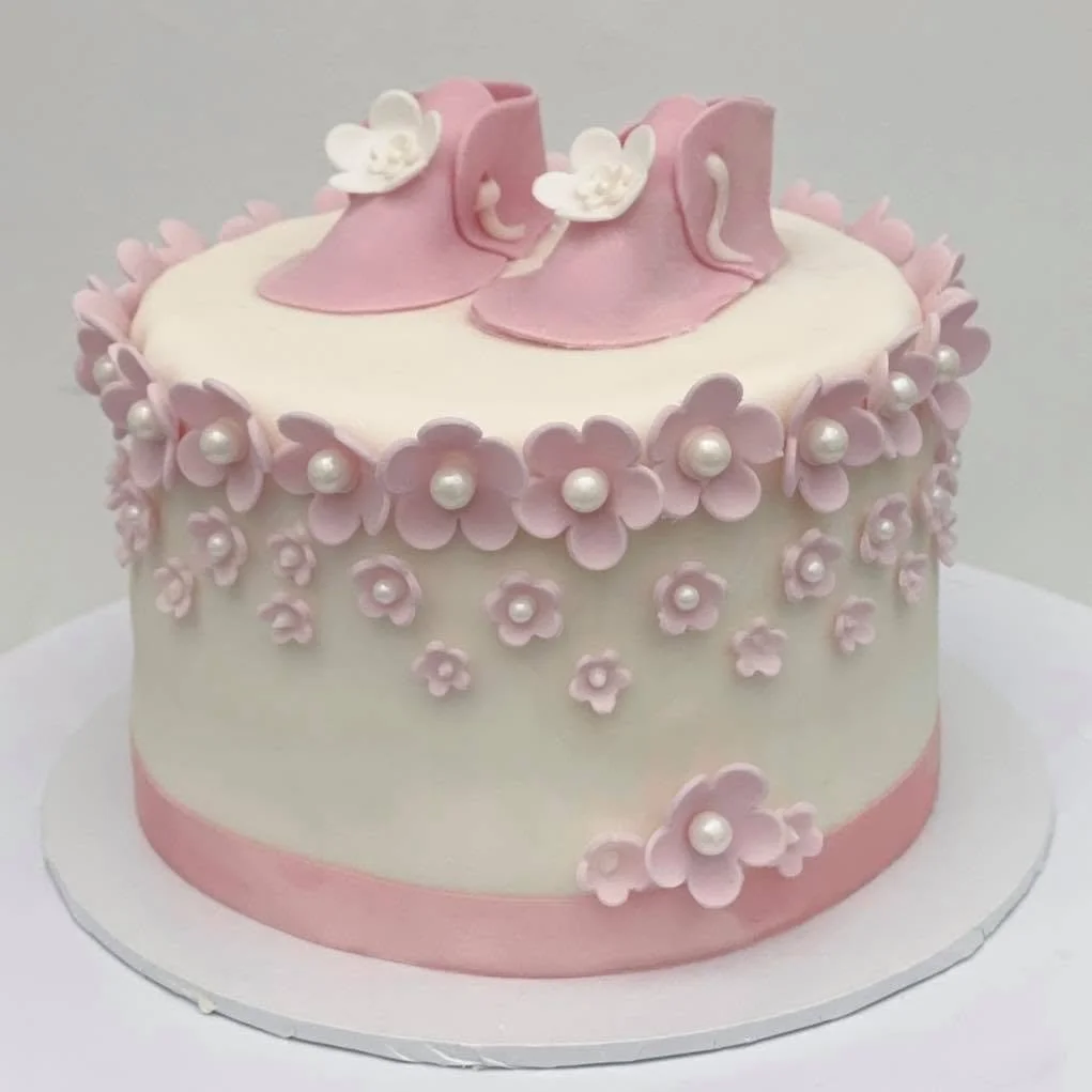 A round cake decorated with pink and white flowers, pearls, and pink shoes on top.