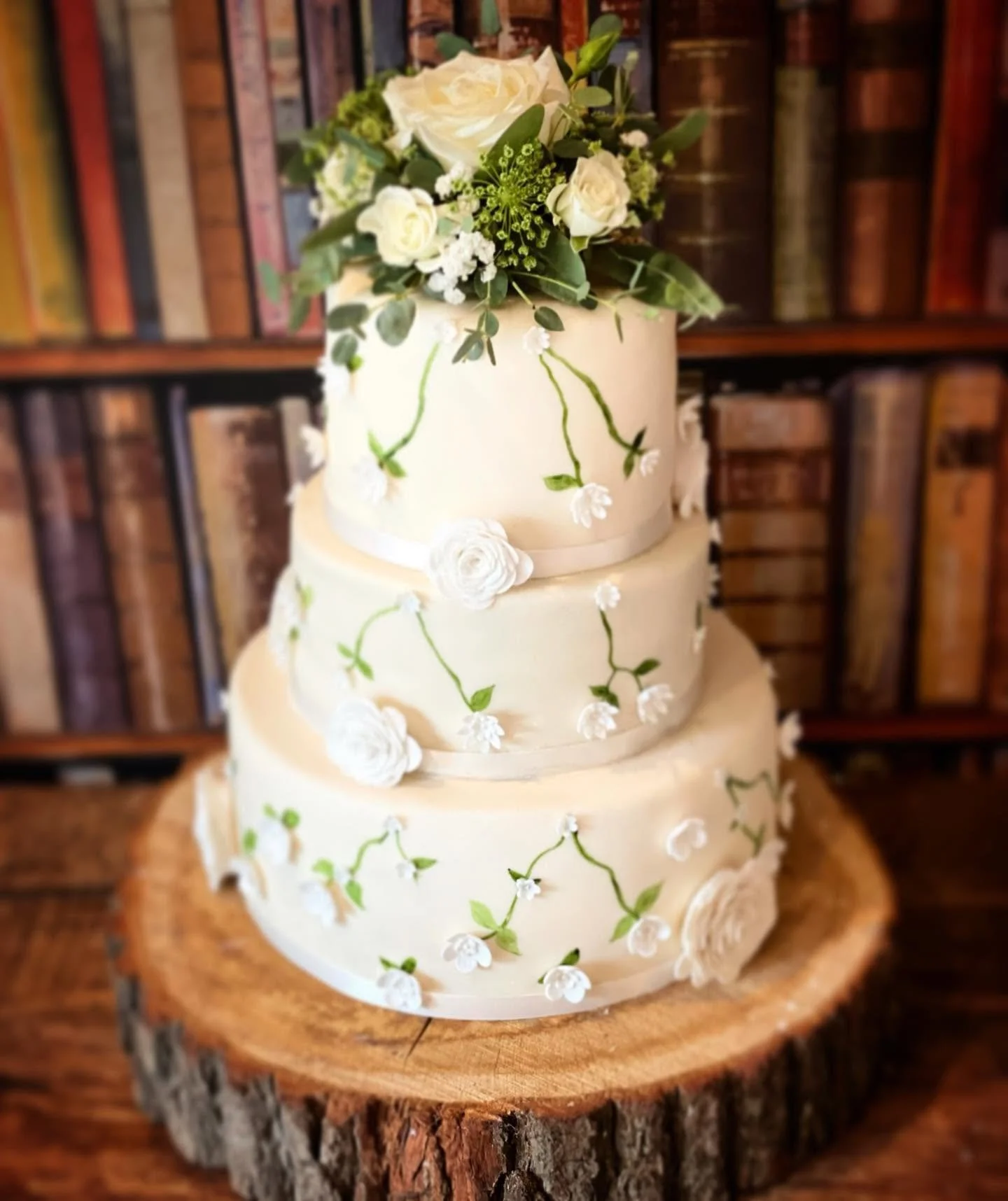 A three-tiered white wedding cake decorated with white roses, small white flowers, and green vines, placed on a wooden slab in front of a bookshelf.