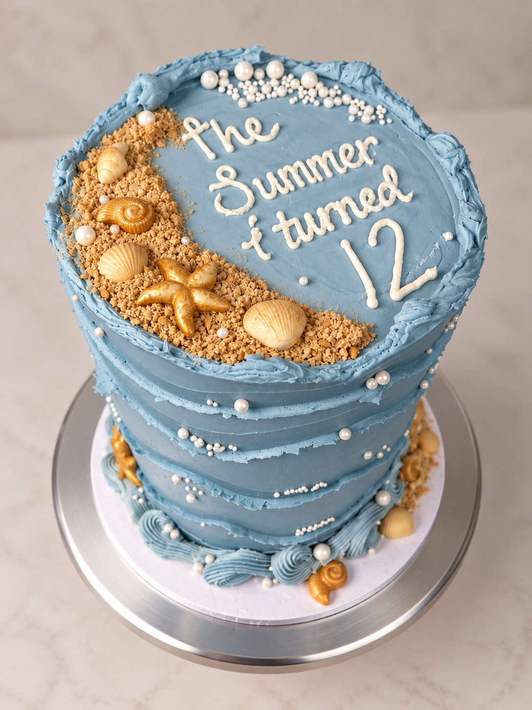 A blue layered birthday cake with white and gold seashell and starfish decorations, white pearl-like beads, and a message written in white icing that says 'the summer i turned 12'.