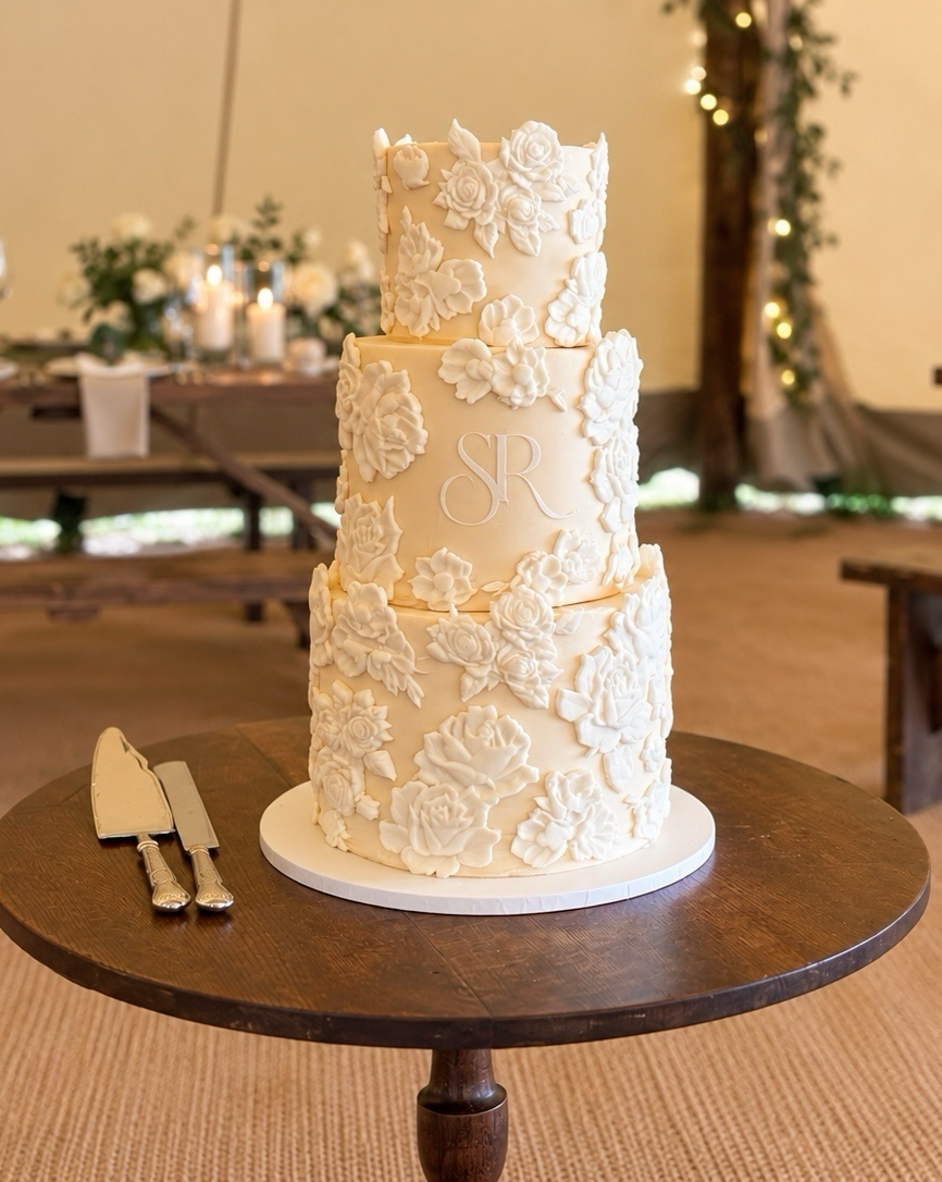 Three-tiered wedding cake with white floral decorations and monogram 'SR' in the center, placed on a dark wooden table with cake knives beside it. Blurred floral arrangements and candles in the background.