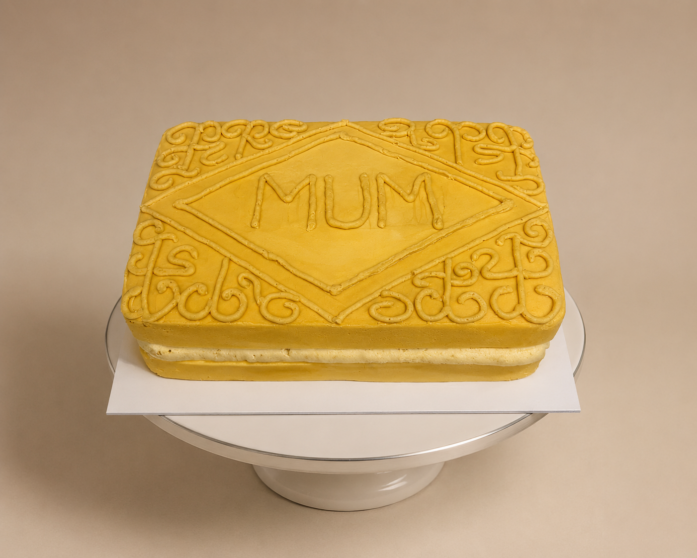 A rectangular cake with yellow icing, decorated with gold piping, displaying the word 'MUM' inside a diamond shape in the center, placed on a white cake board.