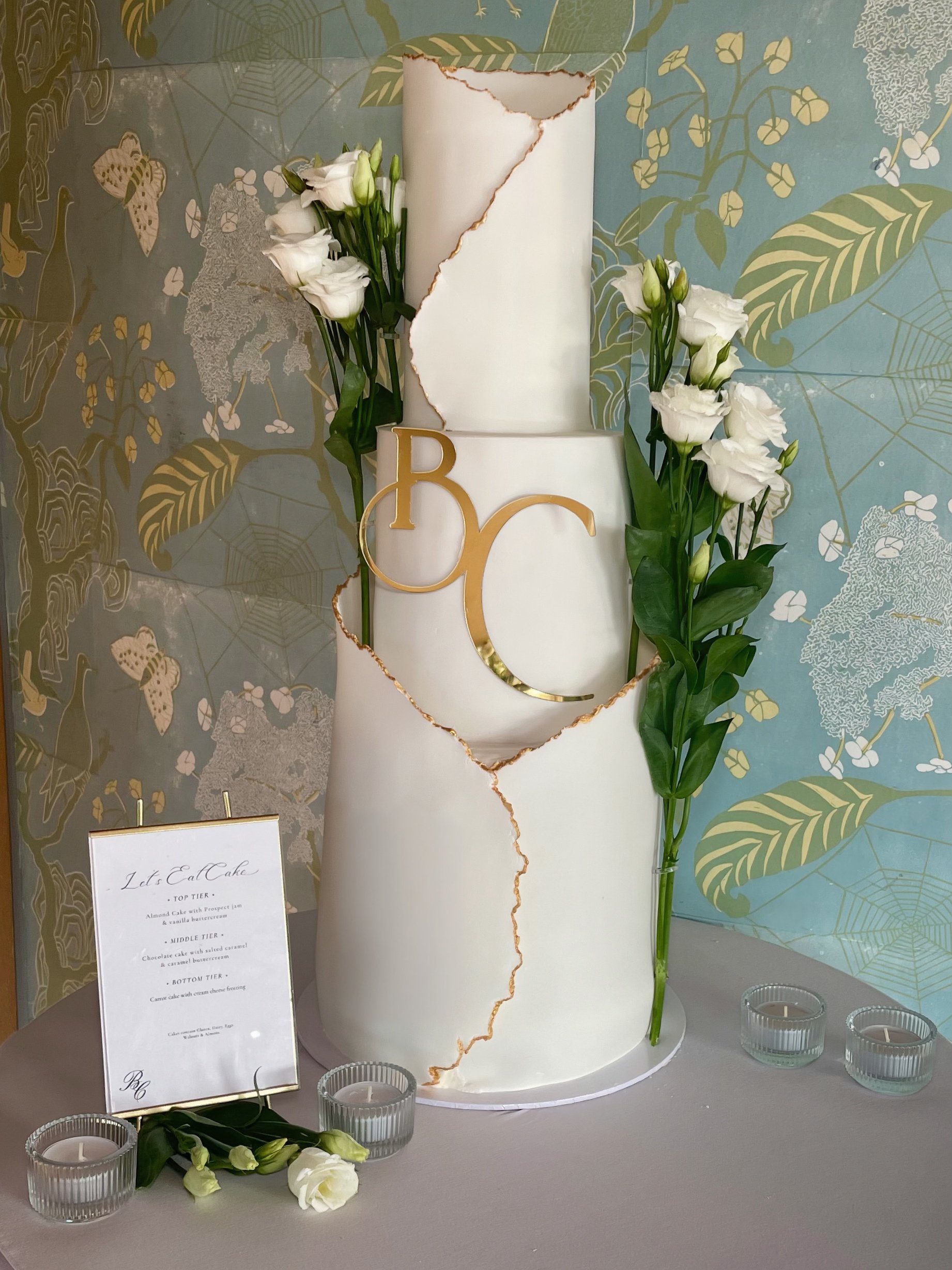 A wedding cake with a large number one topper, decorated with white flowers and greenery, and gold accents in the shape of the letter 'B'. The cake is on a table with candles and a menu card nearby, and a decorative background with butterflies, spider webs, and leaves.