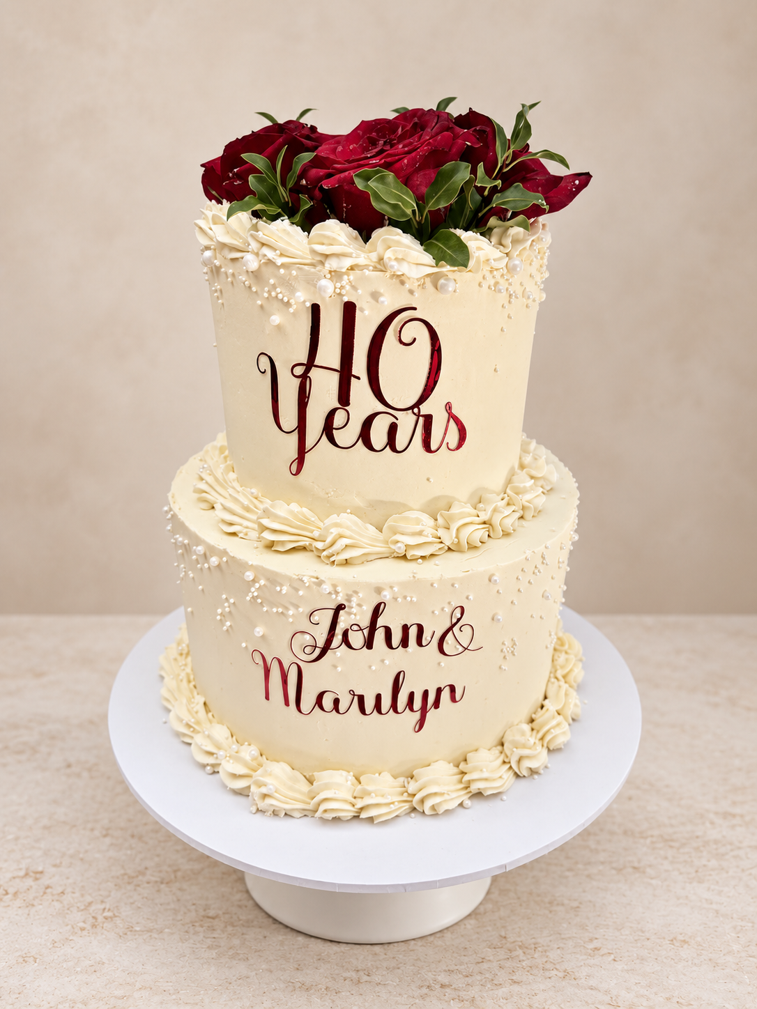 Two-tier wedding cake with white frosting, decorated with cream-colored piping and edible pearls. The top tier is topped with red roses and green leaves. Ruby wedding anniversay