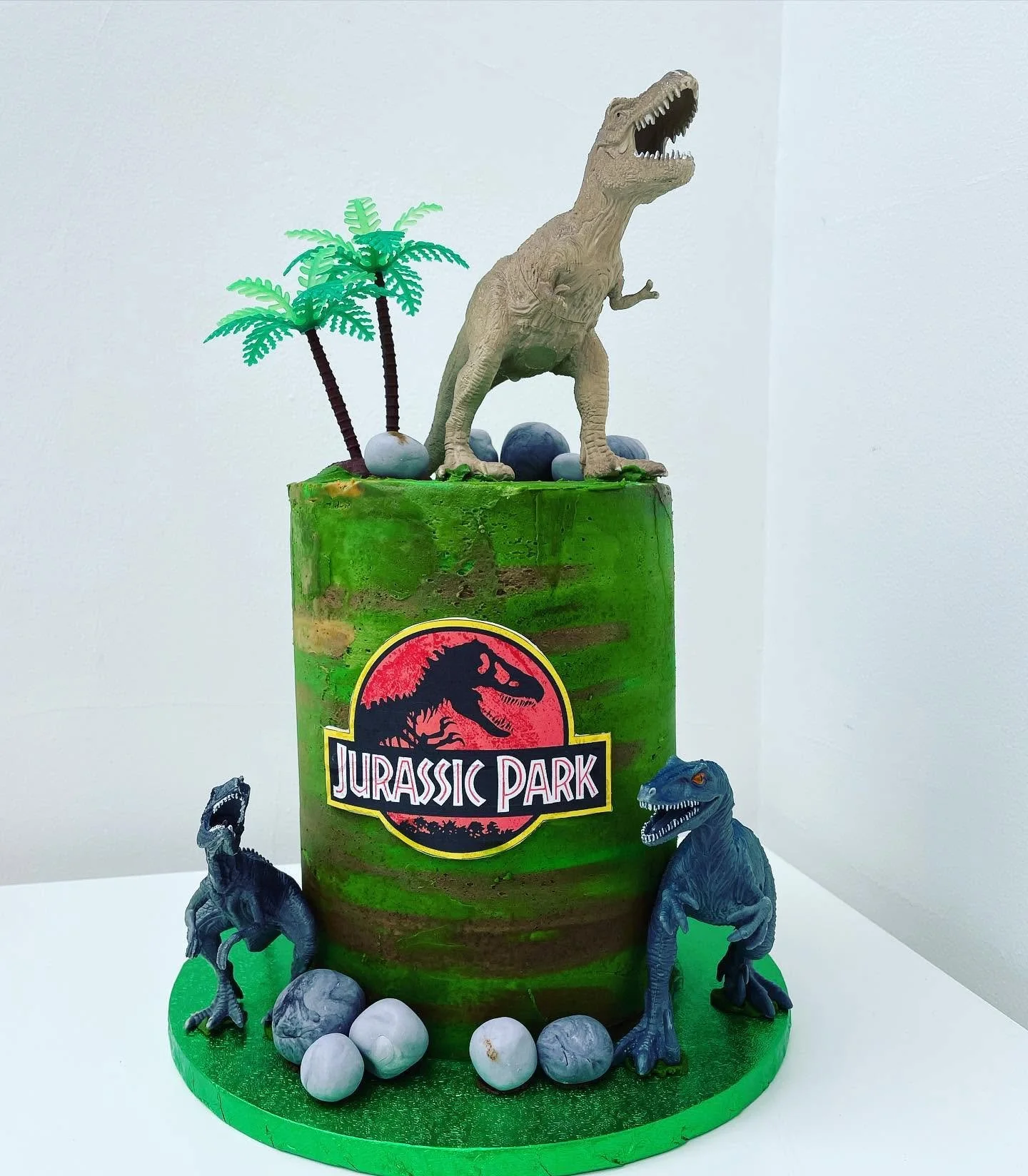 A themed birthday cake designed to resemble Jurassic Park. The cake has two tiers with a green and brown color scheme, decorated with toy dinosaurs, palm trees, and rocks. The upper tier features a large dinosaur figure with an open mouth, and the front displays the Jurassic Park logo.