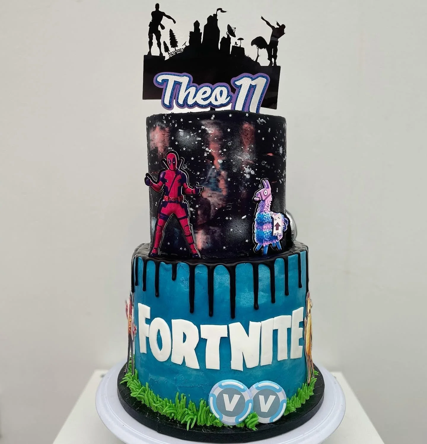 A three-tier birthday cake decorated with a Fortnite theme, featuring Fortnite characters and gaming elements, with a black and blue color scheme, topped with a superhero figurine silhouette and a