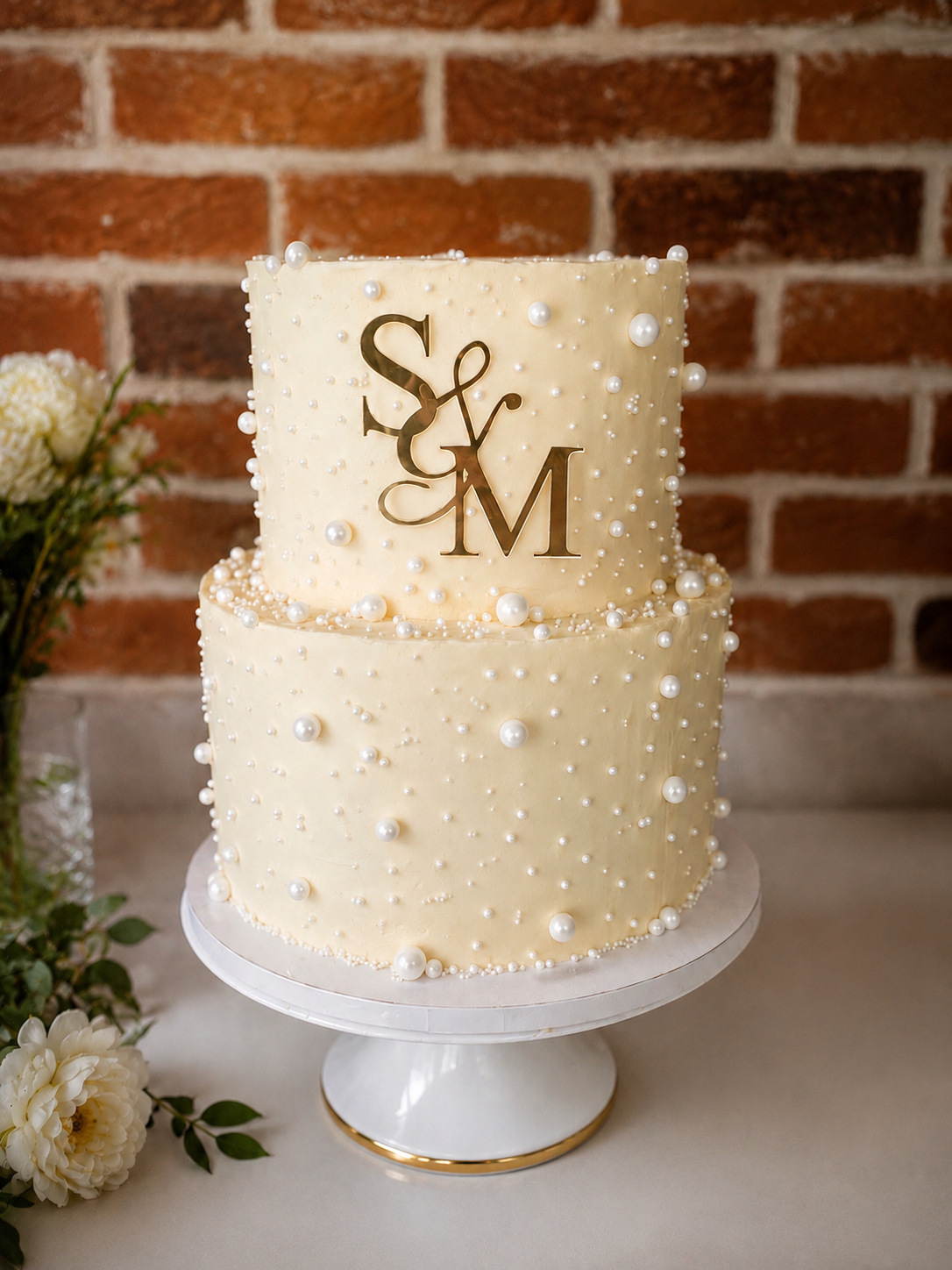 Two-tier cream-colored wedding cake decorated with white pearls, with gold initials 'S & M' on top, placed on a white cake stand, with flowers and a brick wall background.