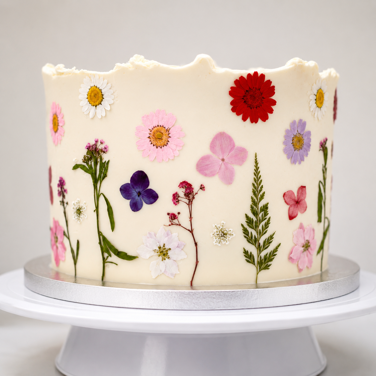 White cake decorated with real pressed flowers in pink, purple, red, white, and yellow, on a silver and white cake stand.