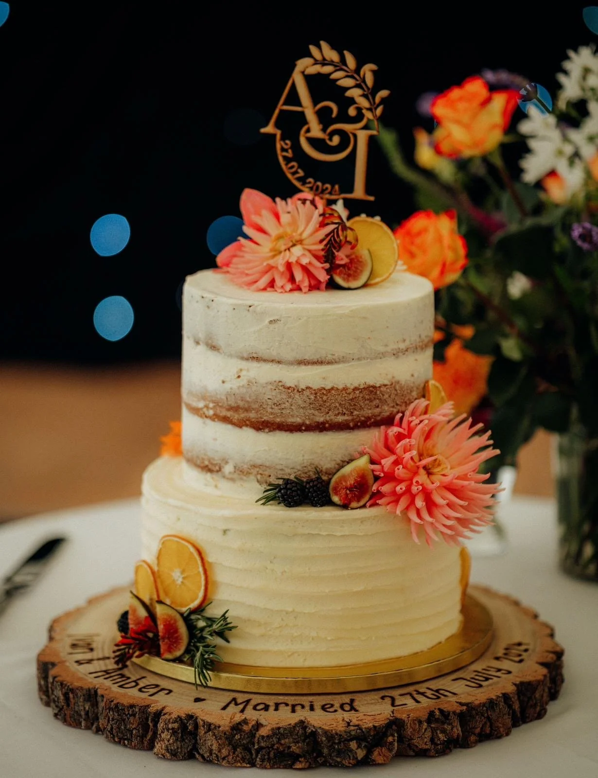 A two-tier wedding cake decorated with pink and orange flowers, figs, blackberries, and citrus slices, with a gold cake topper on a wooden slab engraved with wedding details, and a floral arrangement in the background.