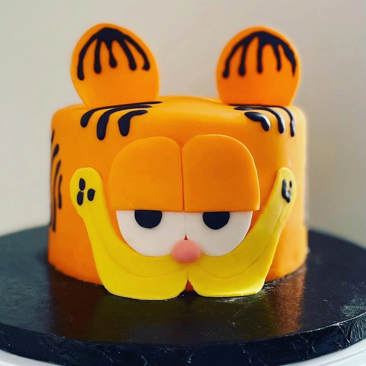 Cake decorated as a cartoon tiger with orange fondant, black stripes, large eyes, and yellow features, placed on a black cake board.
