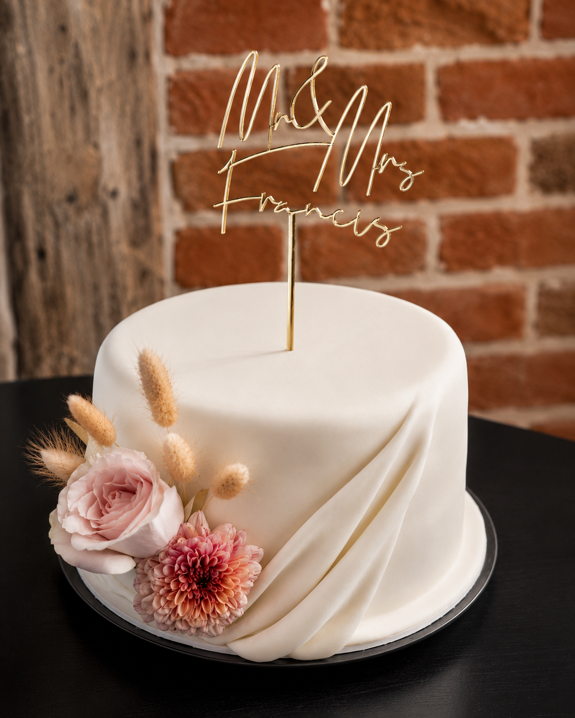 A white wedding cake decorated with pink flowers and a gold topper that says 'Mr & Mrs Frances' against a brick wall background.