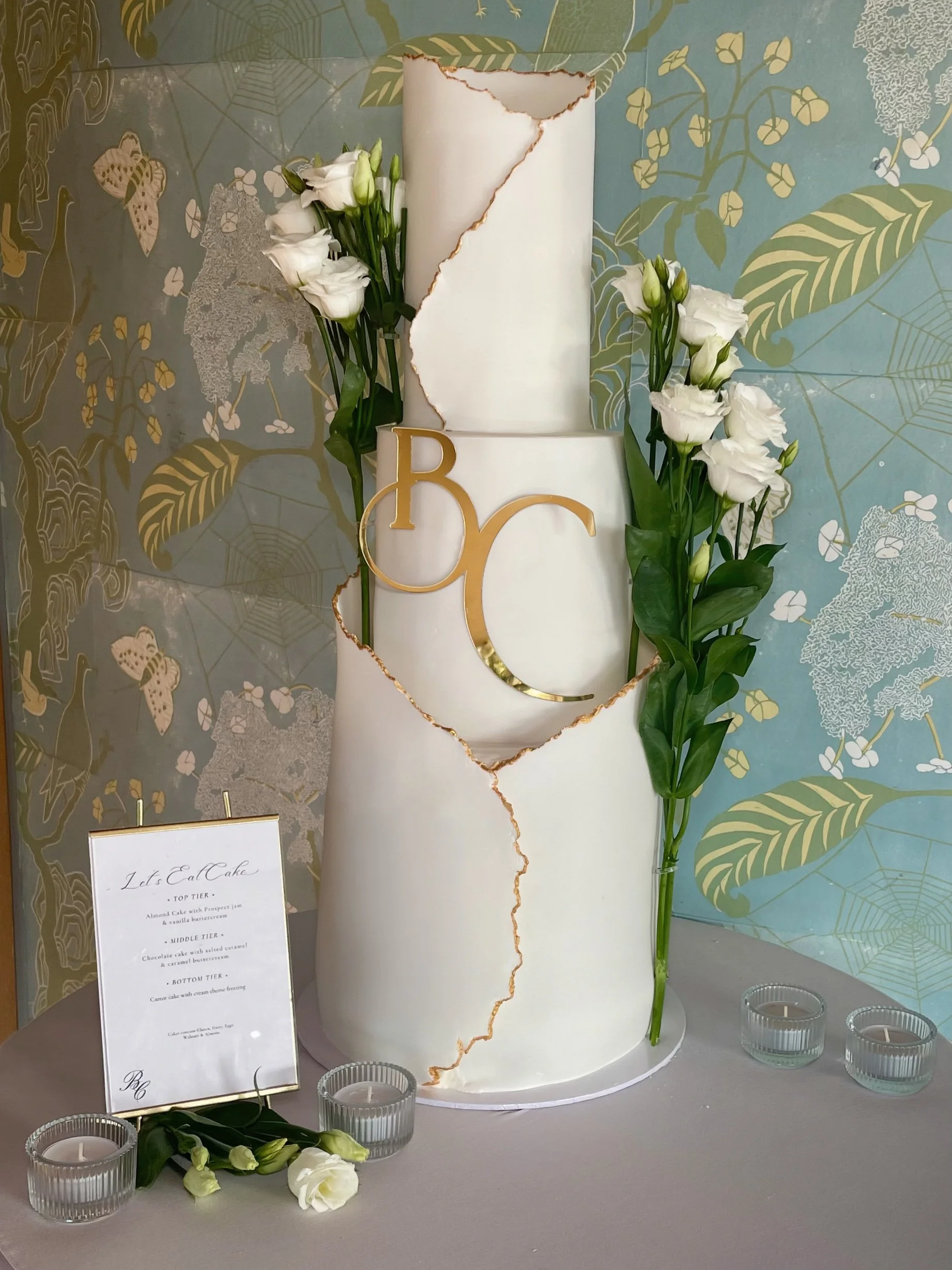 A tall white tiered cake with a torn paper effect revealing gold edges. The cake is decorated with white flowers and is surrounded by small candles and a menu card. The background features wallpaper with green and gold foliage, and there are small glass holders with candles at the front.