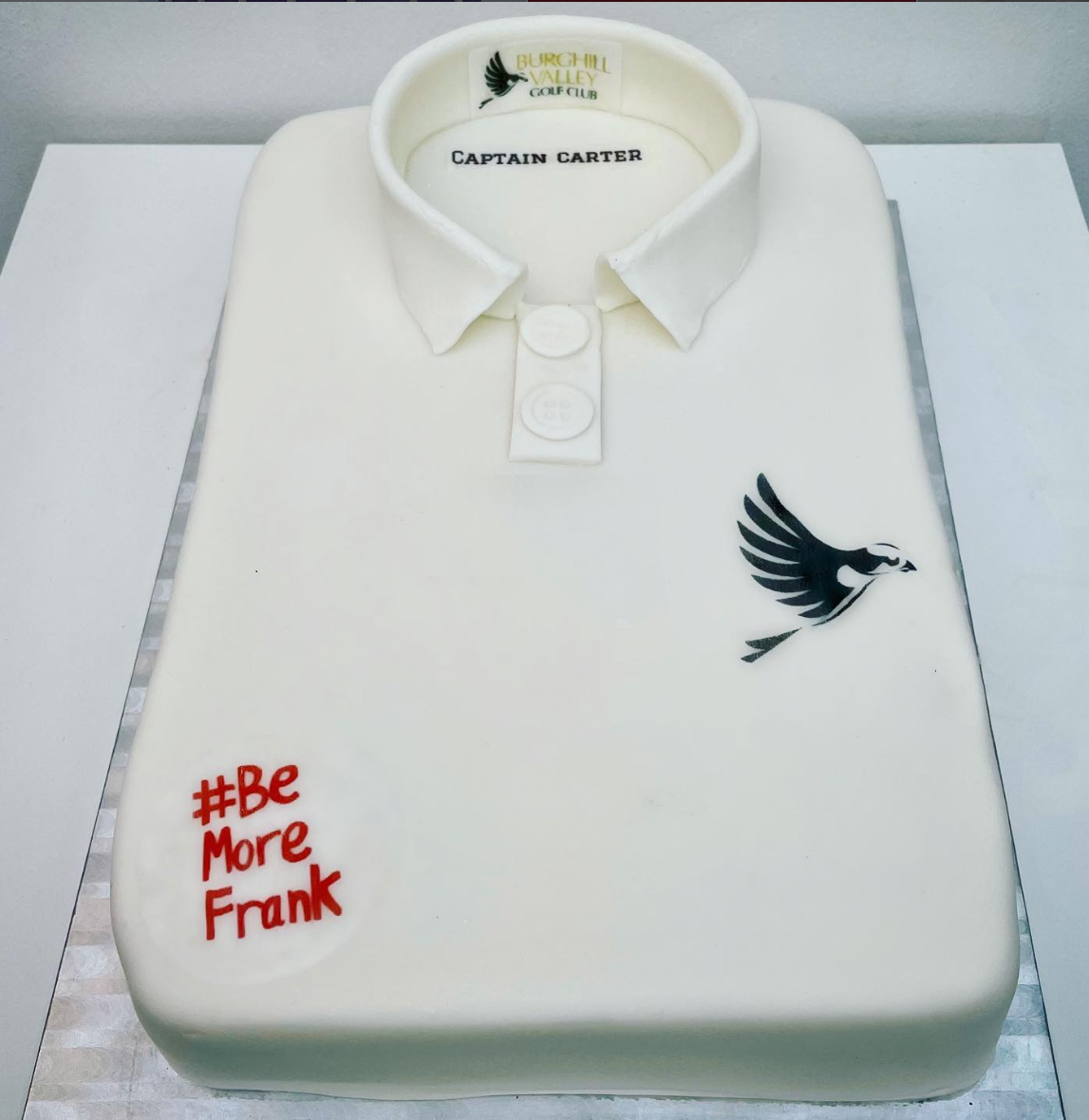 A cake made to look like a white golf shirt with a collar, buttons, and a logo from Burchell Valley Golf Club. The cake also features a black and white bird illustration and the hashtag '#Be More Frank' written in red.