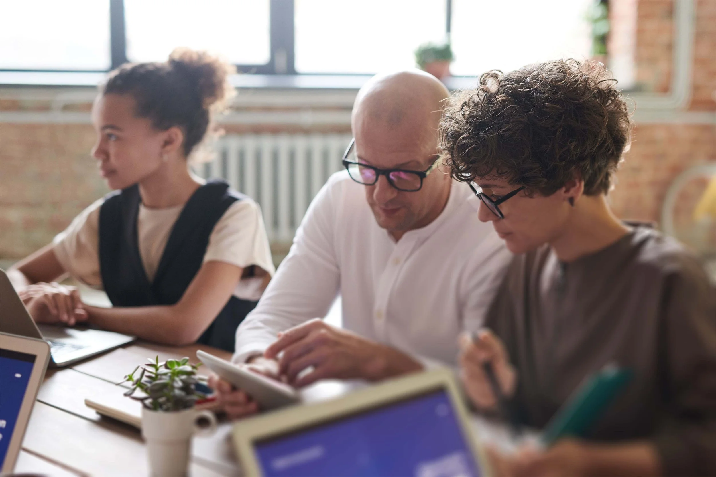 Three people sitting at a table, working or studying, with laptops and notebooks in a bright room with large windows and brick walls.