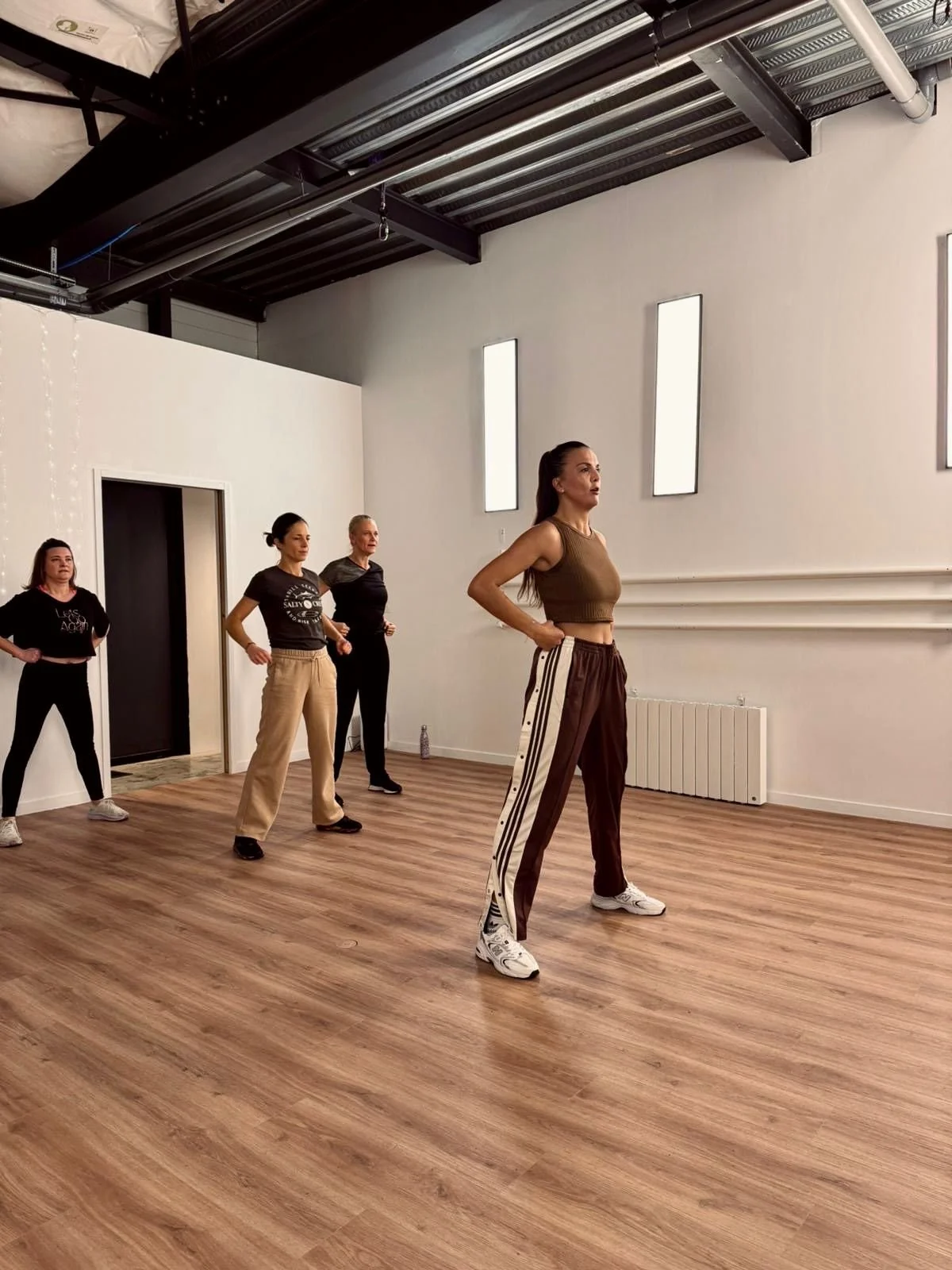 Quatre femmes dansant dans un studio de danse avec un sol en bois et un plafond industriel. La femme à l'avant porte un pantalon de survêtement marron clair et un crop top marron. Les autres femmes portent des vêtements décontractés noirs ou beige.