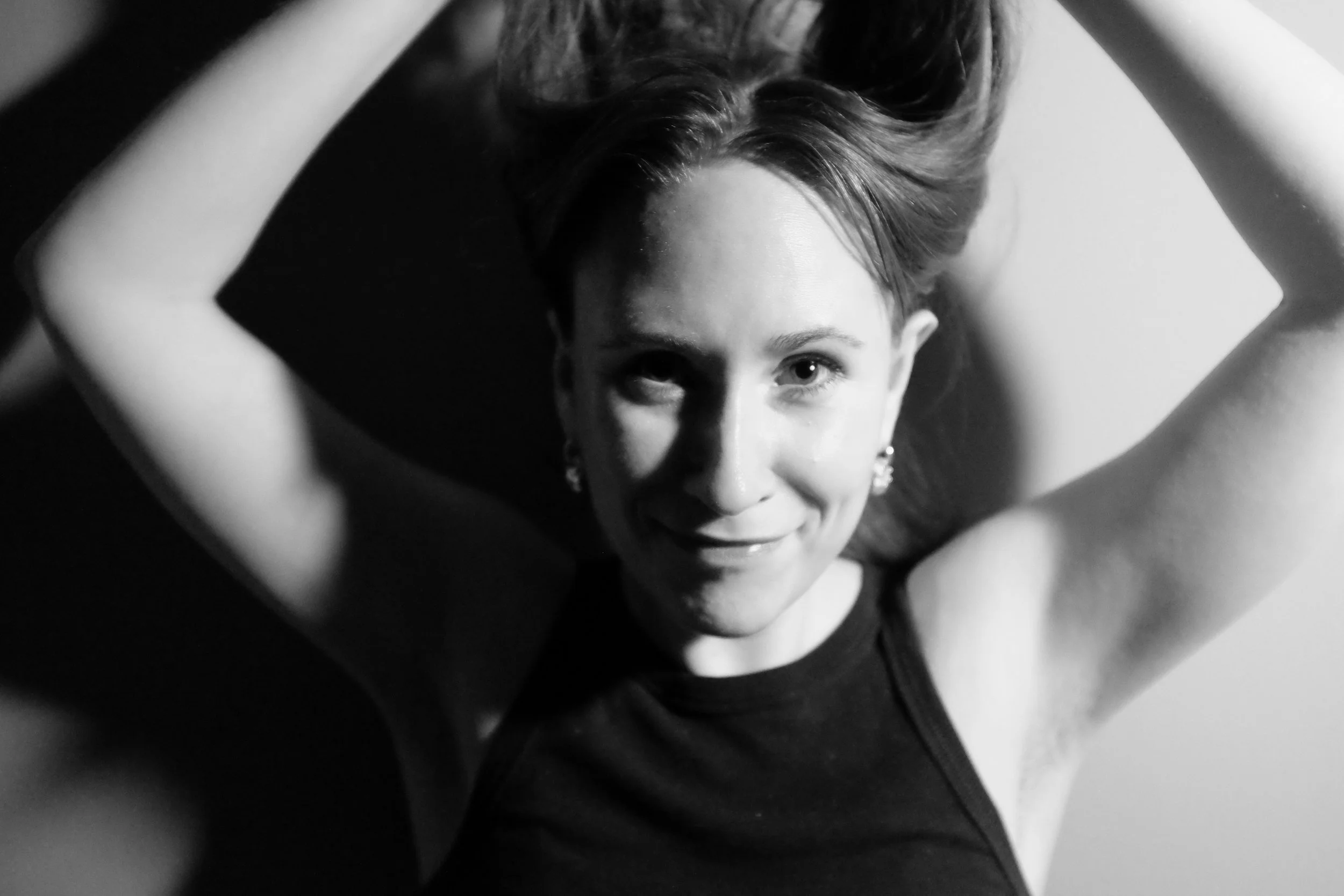 Black and white photo of a woman with shoulder-length hair, smiling and looking directly at the camera, with arms raised and hands behind her head.