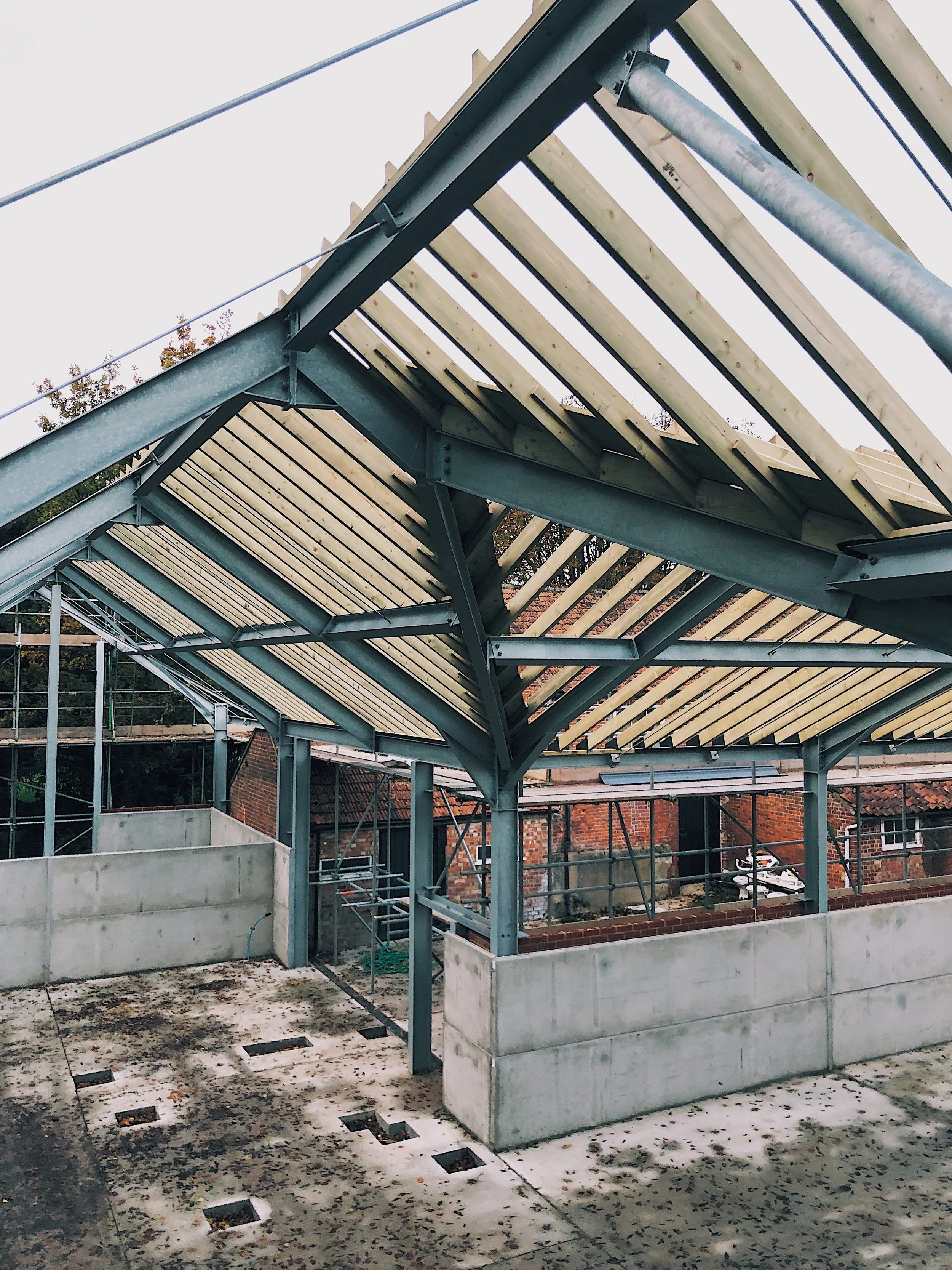 Construction site showing the framework of a building with metal beams and wooden slats, concrete foundation, and scattered leaves on the ground.