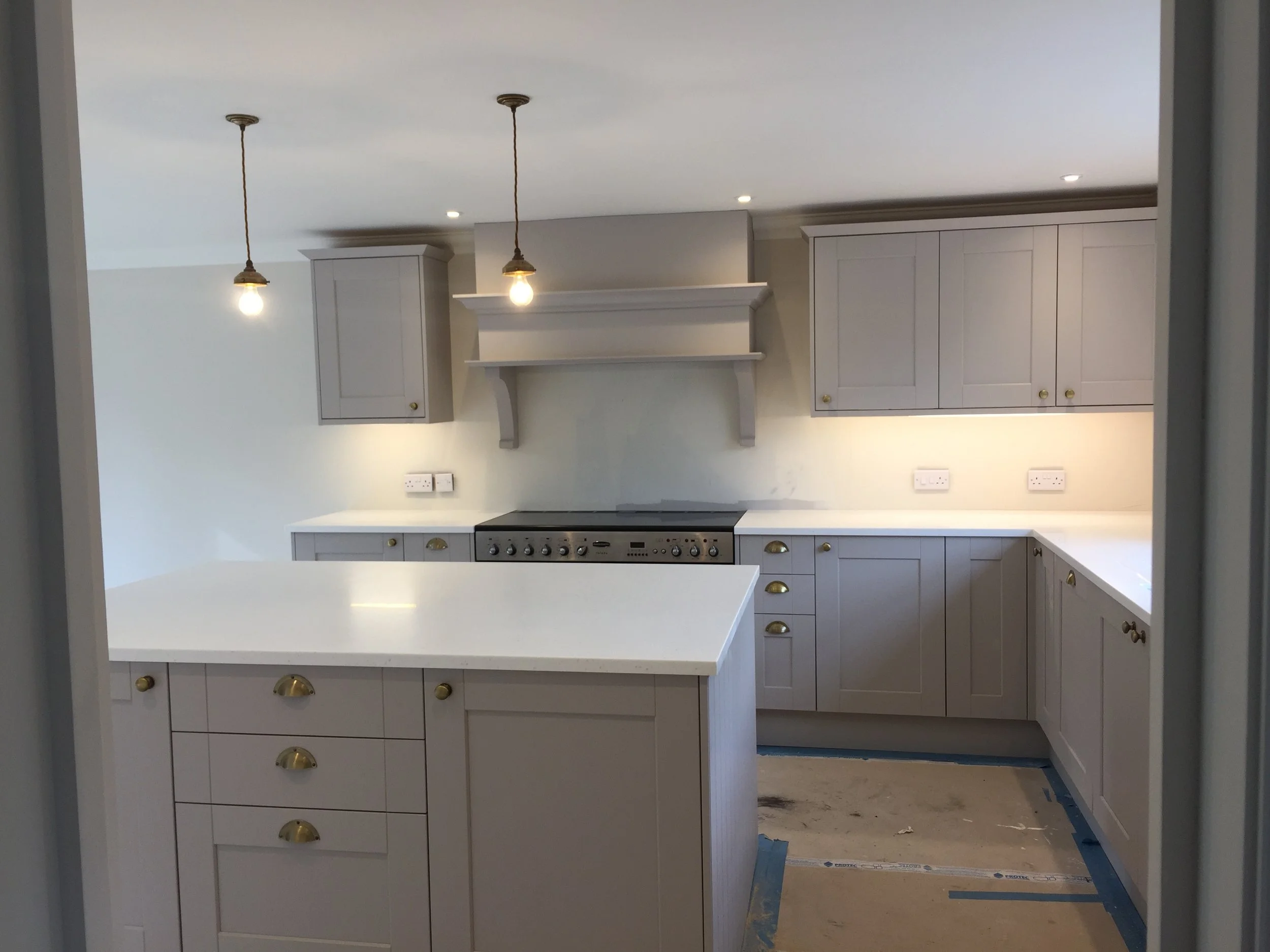 A kitchen under construction with white cabinetry, a stovetop, and pendant lighting hanging from the ceiling. The area appears to be in the process of being finished with some exposed flooring and construction materials visible.