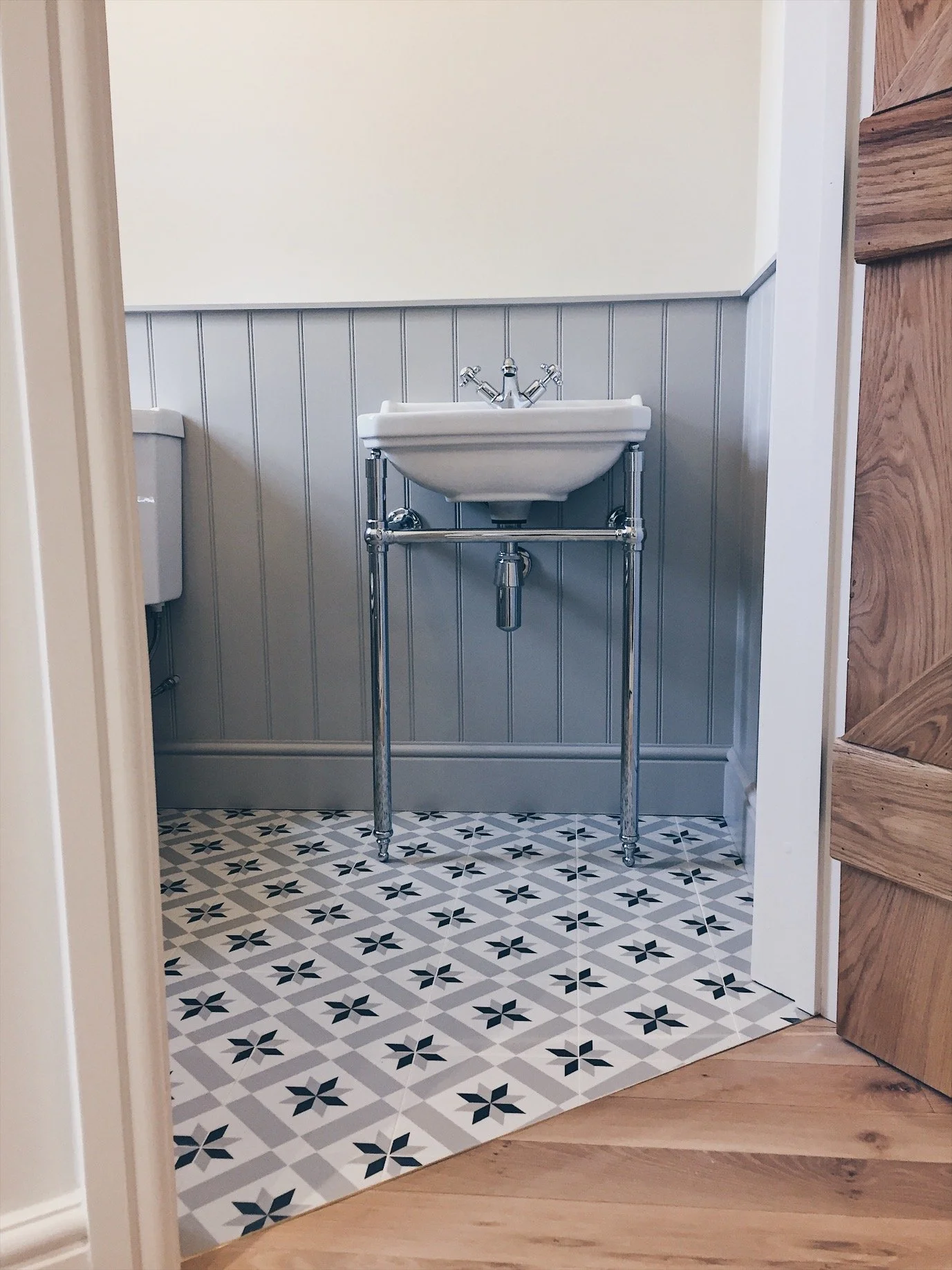 A laundry room with a vintage white ceramic utility sink supported by chrome legs against a wall with gray vertical shiplap paneling. The floor is a patterned tile with black star-like designs on a white background, adjacent to a wood floor.