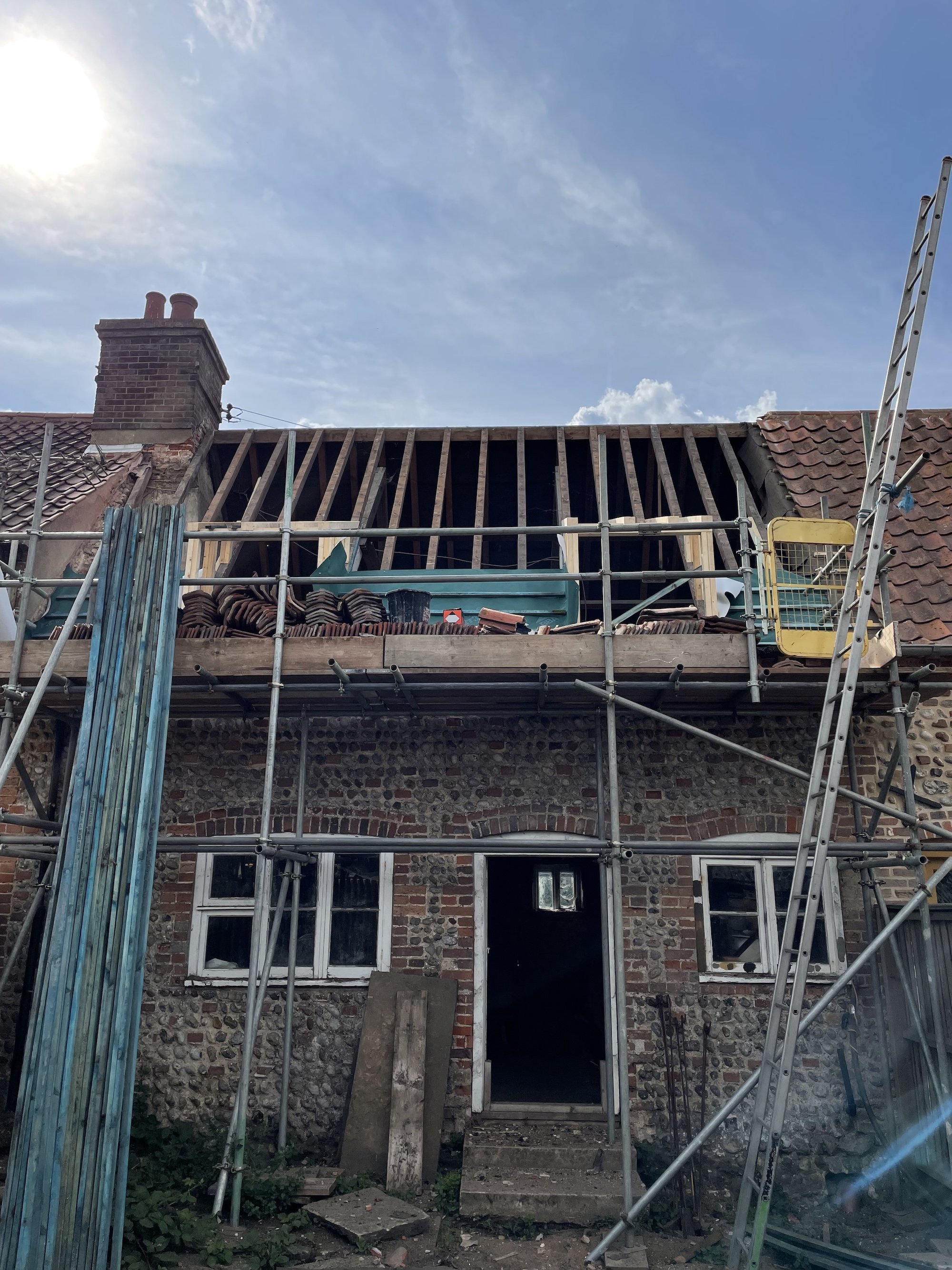 The upper part of a house under renovation with exposed roof rafters and a partially removed roof. There is scaffolding in front of the house, and a TV antenna is visible on the roof. Parts of the roof tiles are stacked on the scaffolding. The buildi